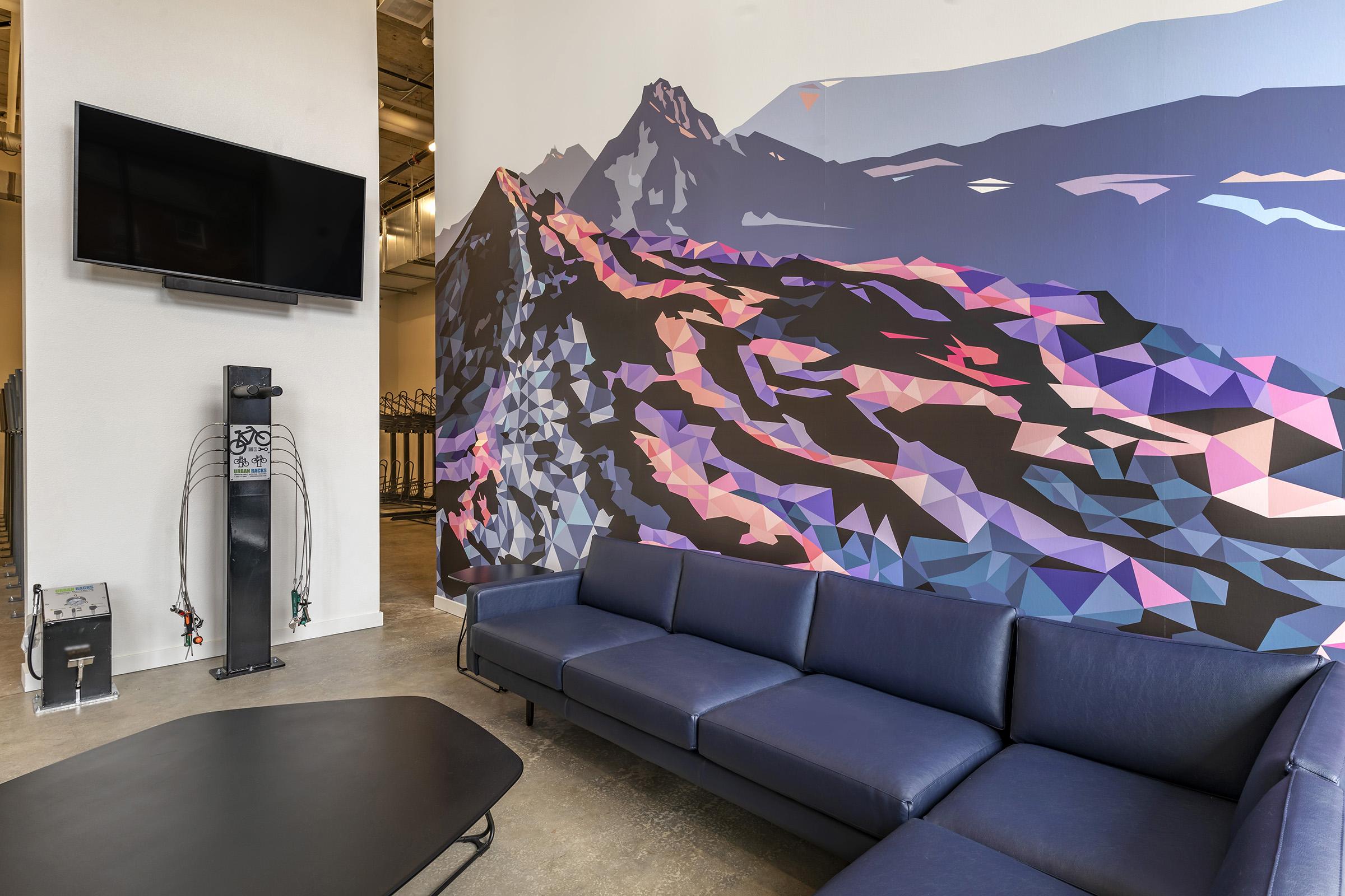 A modern lounge area featuring a blue sectional sofa and a large geometric mural of a mountain landscape on the wall. A black coffee table is placed in front of the sofa, and a flat-screen TV is mounted above. The space has an industrial feel with concrete flooring and contemporary decor elements.