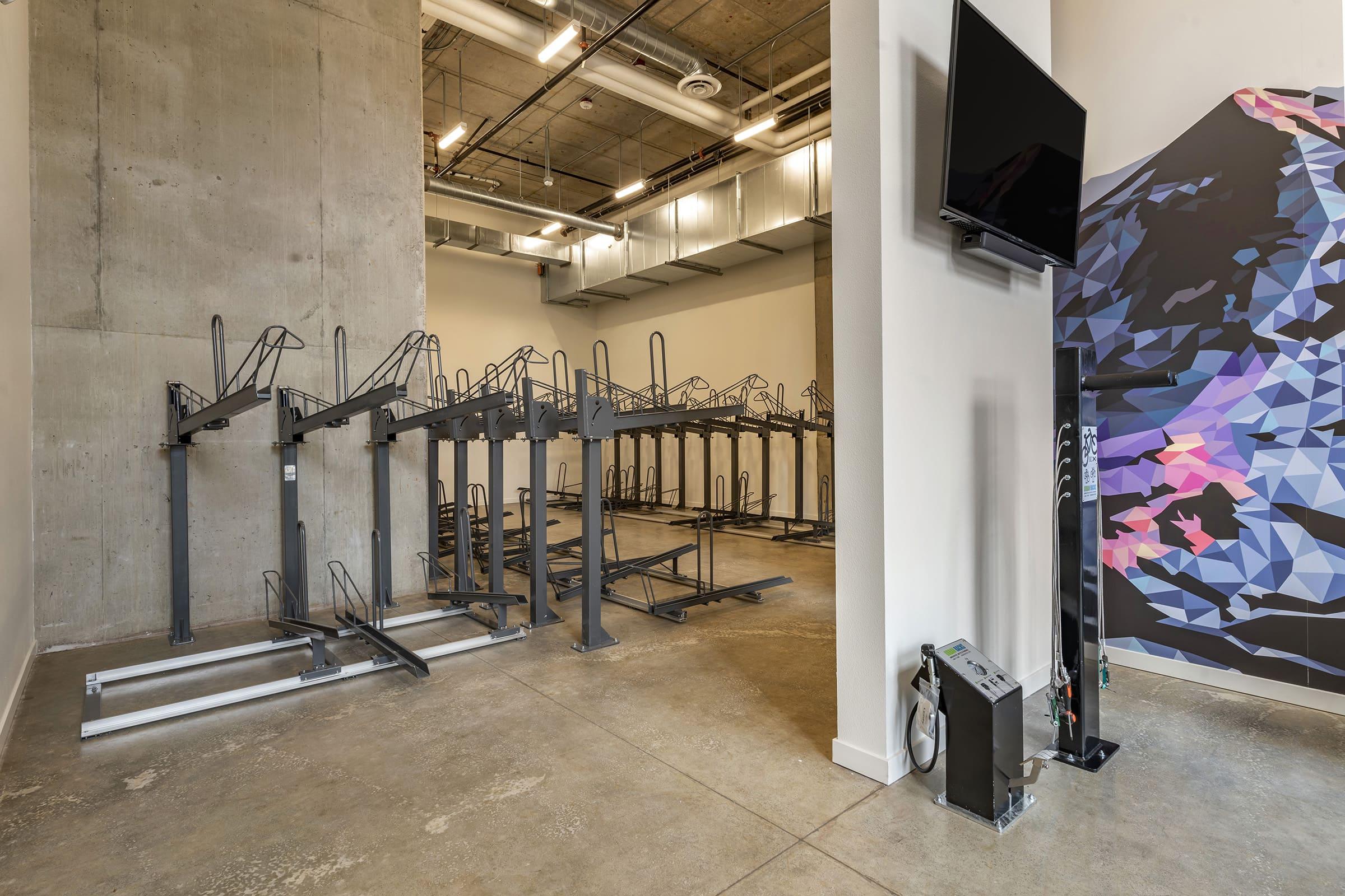Interior of a modern room featuring bike racks against a concrete wall, with a large flat-screen TV mounted on a wall. The space is well-lit and has a contemporary design, with minimalistic elements and a geometric wall art design in the background.