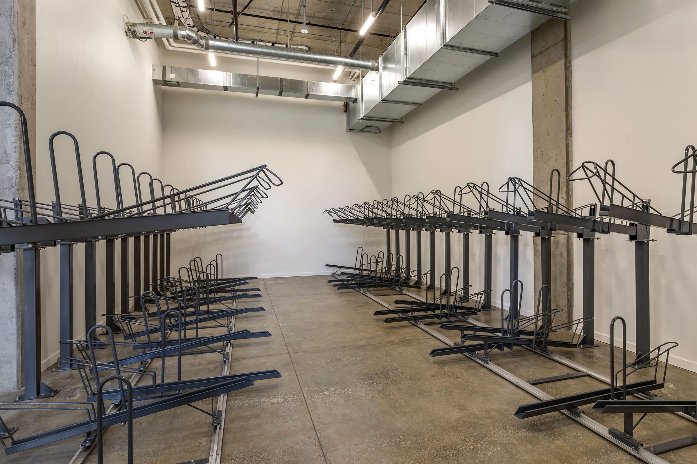 A spacious room with a concrete floor and white walls, featuring multiple bike racks arranged in parallel lines. The racks are metallic and each has a horizontal support, creating an organized layout for bike storage. Natural light enters through large windows, enhancing the minimalist design.