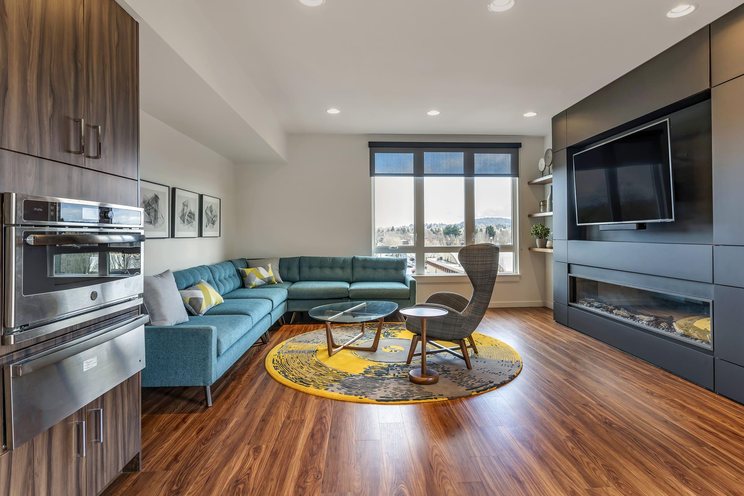 Modern living room featuring a teal sectional sofa, glass coffee table, and a patterned yellow rug. A wall-mounted TV sits above a contemporary electric fireplace. Large windows provide natural light, showcasing a scenic view. Elegant cabinetry and a built-in oven complete the stylish interior design.