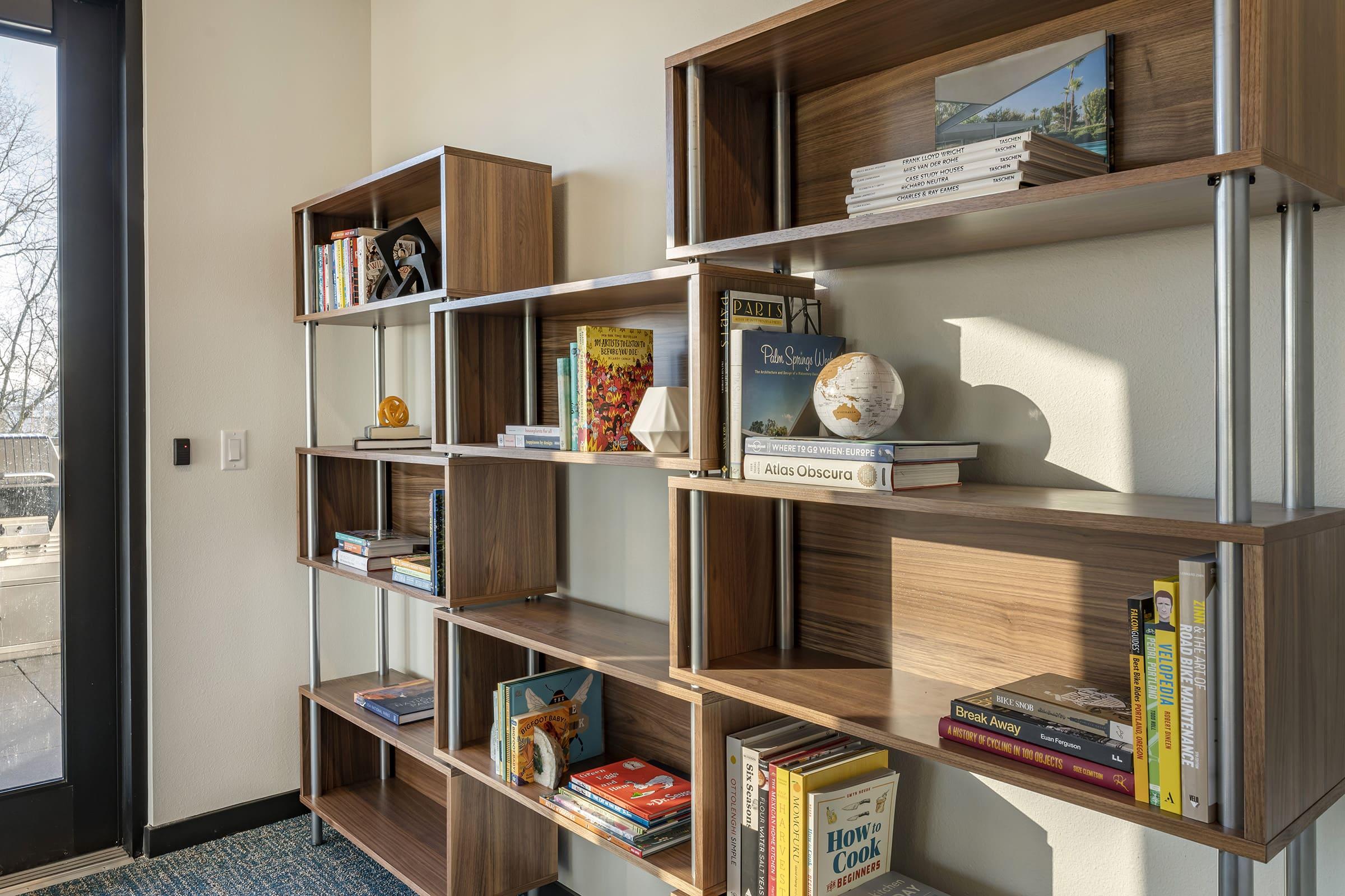 A modern bookshelf with various books, decorative items, and a globe. The shelves are made of wood and metal, displaying a mix of colorful book covers and small decor pieces, set against a neutral wall in a well-lit room.