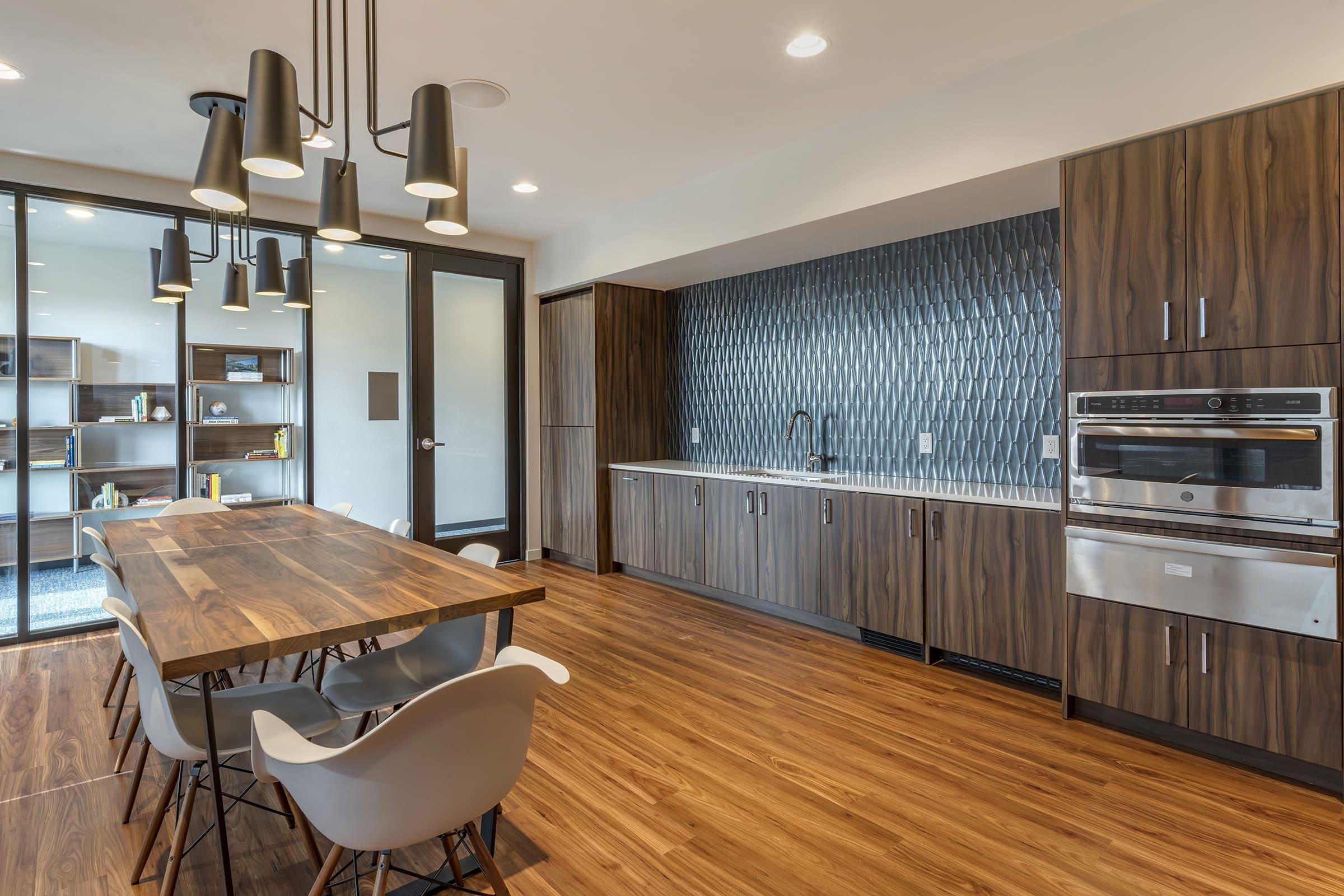 Modern kitchen and dining area featuring a wooden dining table with white chairs, sleek cabinetry, stainless steel appliances, and a textured wall design. The space has large windows allowing natural light, pendant lights above the table, and a contemporary aesthetic with warm wood flooring.
