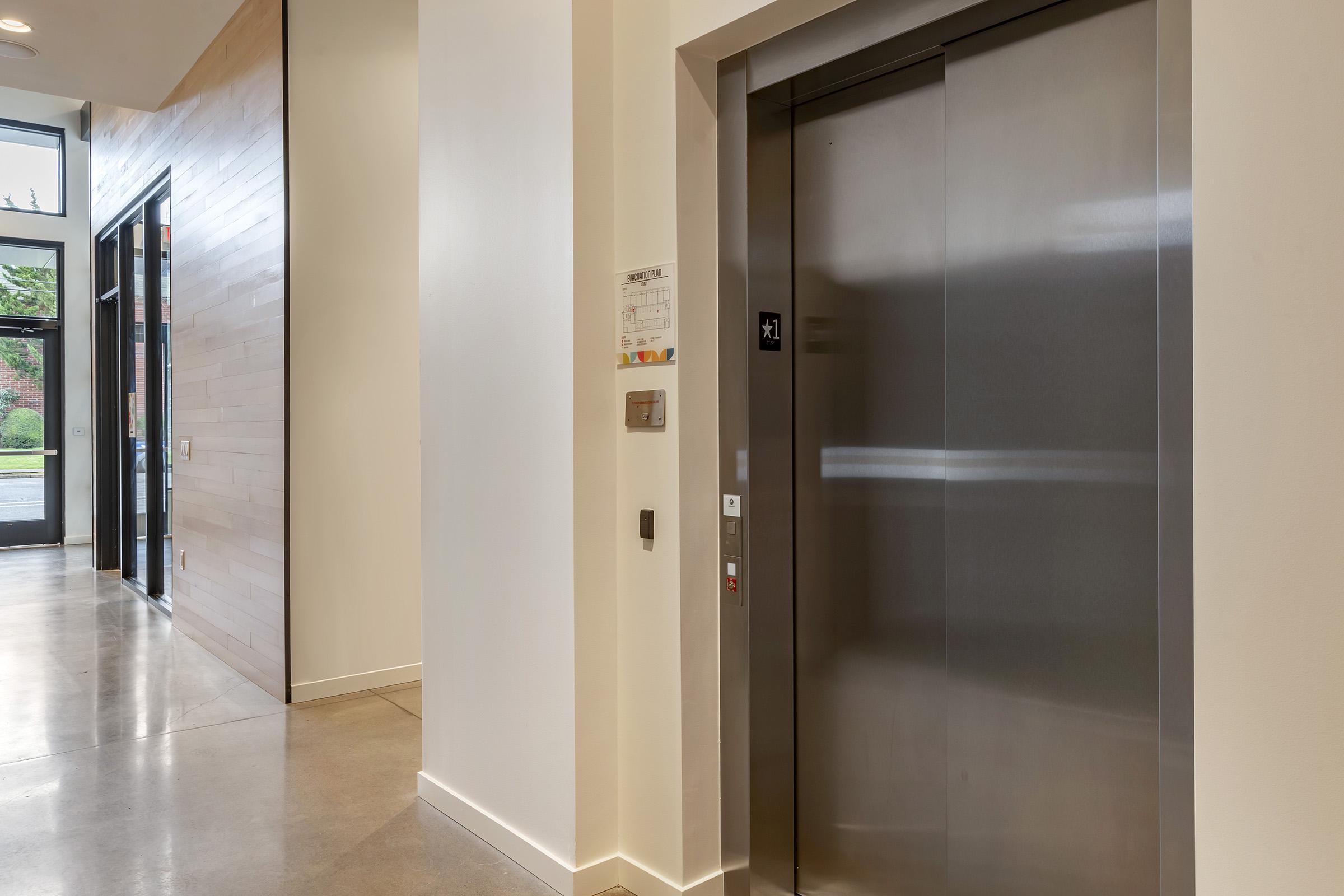 Modern elevator in a spacious hallway with large windows, featuring a minimalist design. The floor is made of polished concrete, and there's a wooden wall paneling. A directory and elevator controls are visible next to the elevator door.