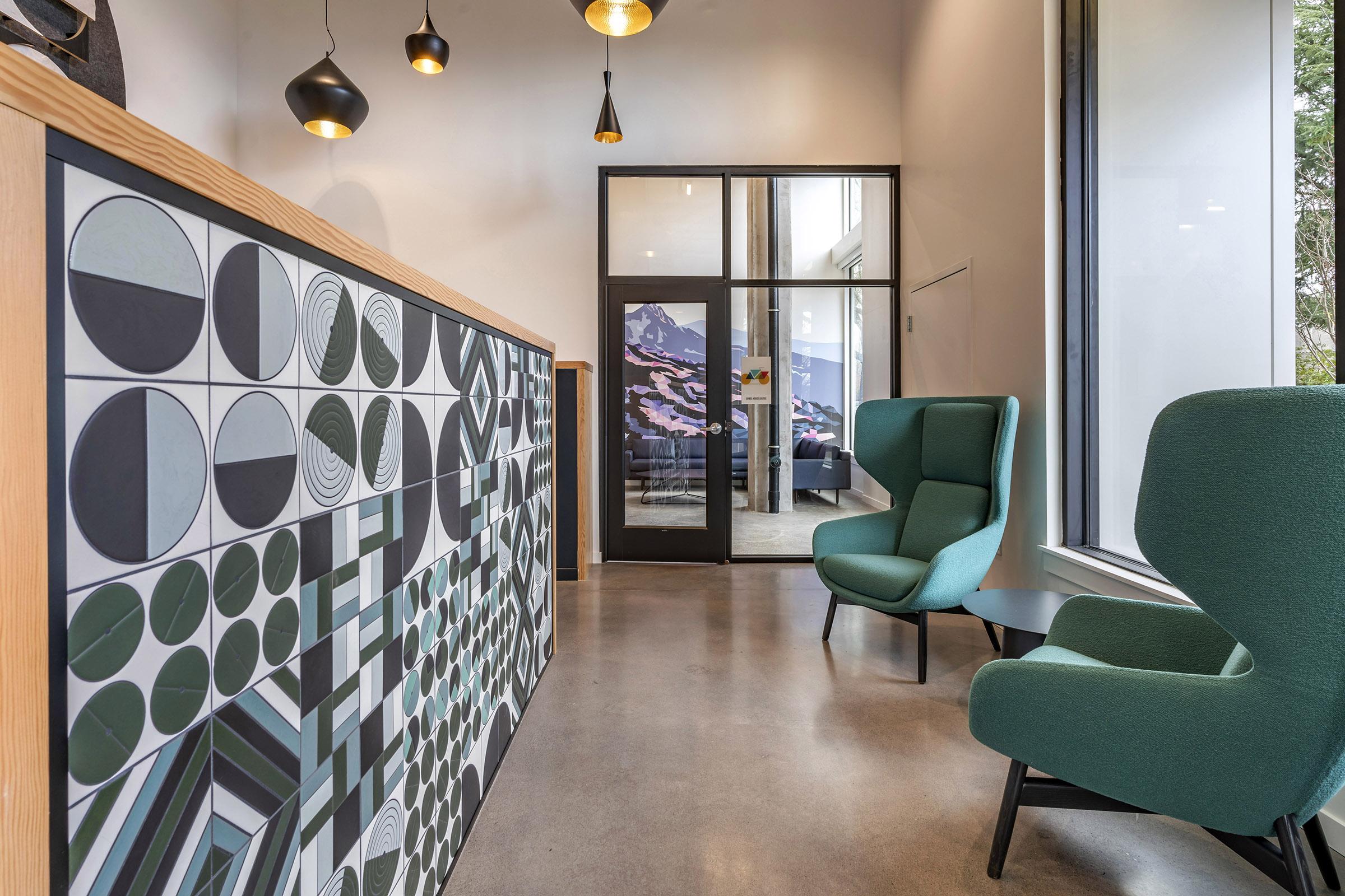 Modern interior of a lobby space featuring a textured wall with geometric patterns in black, white, and green, two green lounge chairs, a coffee table, and contemporary lighting fixtures. Large windows provide natural light and a view of the outdoors.