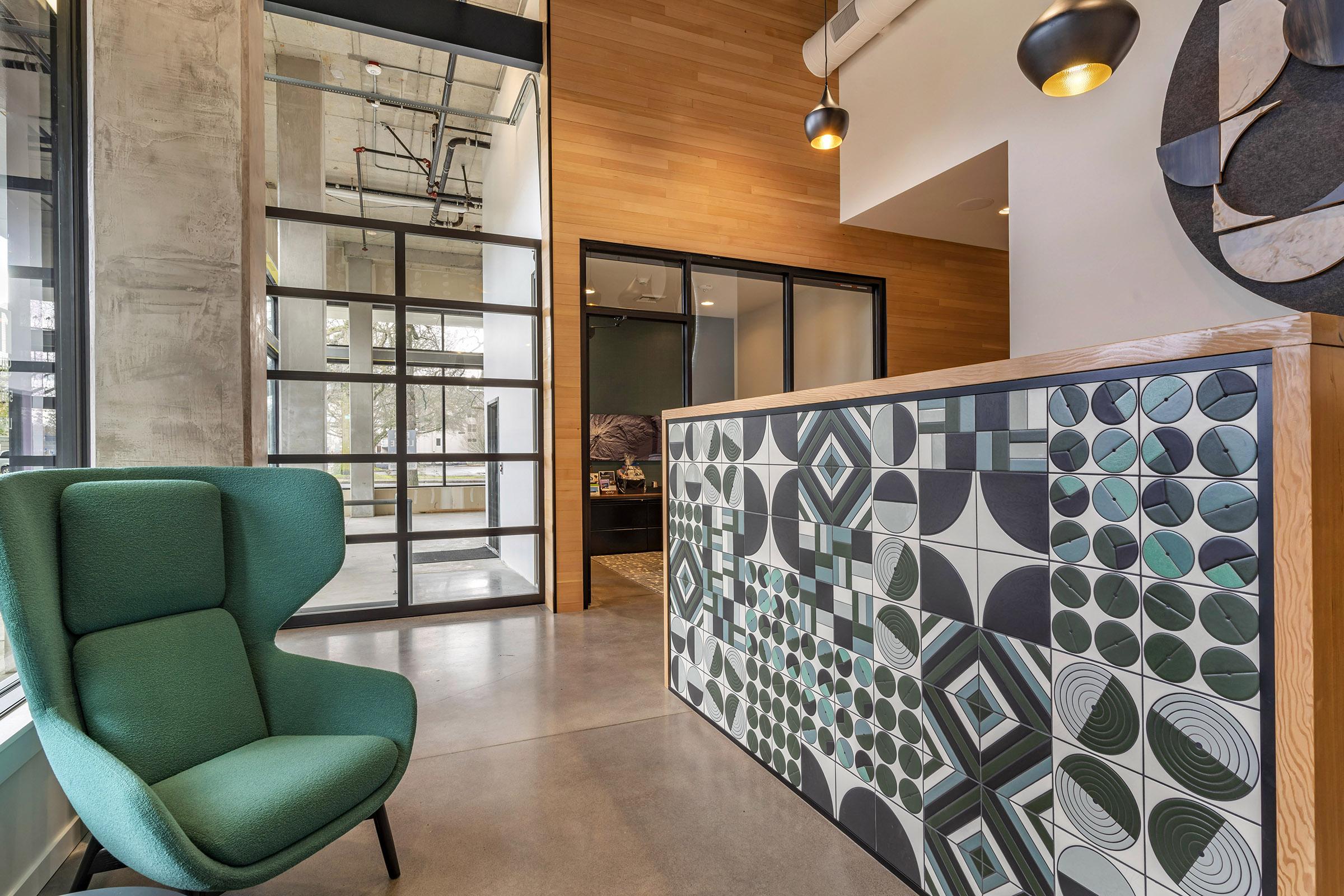 A modern office reception area featuring a geometric-patterned front desk, a green armchair, and large glass doors. The walls are a mix of wood and concrete, with warm lighting provided by pendant lamps. Natural light filters through the windows, creating an inviting atmosphere.