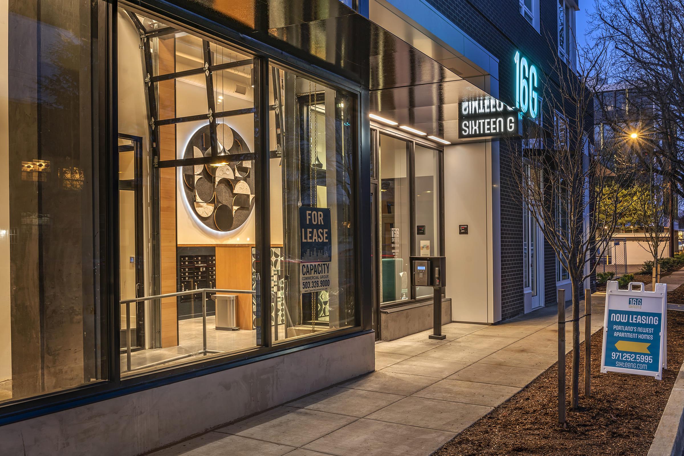 Modern building exterior at night featuring large glass windows. A sign indicates “FOR LEASE” with contact information. The entrance displays stylish architectural elements, including circular decorations and ambient lighting. A leasing sign is located in front, advertising available units.