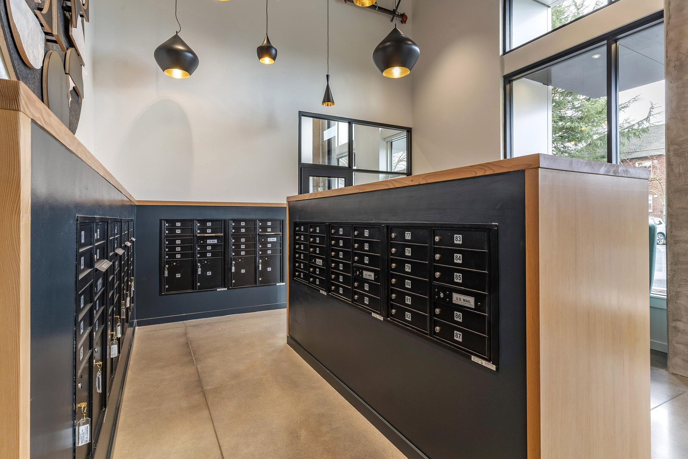 Interior view of a modern postal area featuring a row of black mailboxes against a light-colored wall. The space has large windows allowing natural light, and stylish pendant lights hang from the ceiling. The floor is polished concrete, creating a contemporary atmosphere.