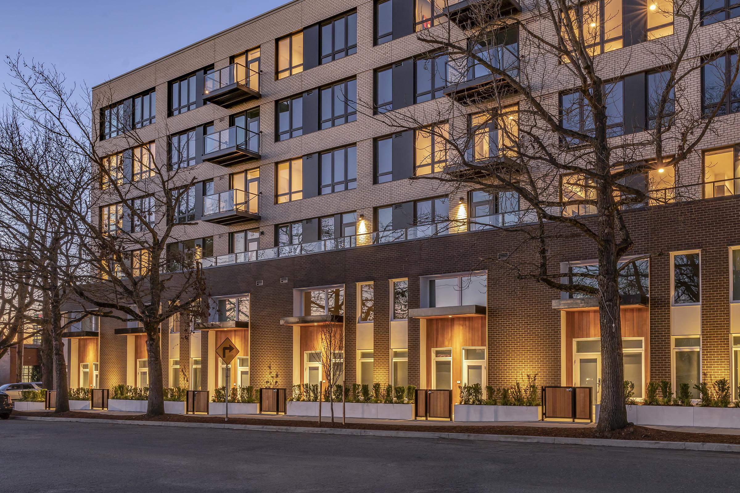 A modern apartment building at dusk, featuring a combination of brick and large glass windows. Balconies are visible on the upper floors, and trees line the street in front. Soft lighting from the windows highlights the architectural details, creating a warm and inviting atmosphere.