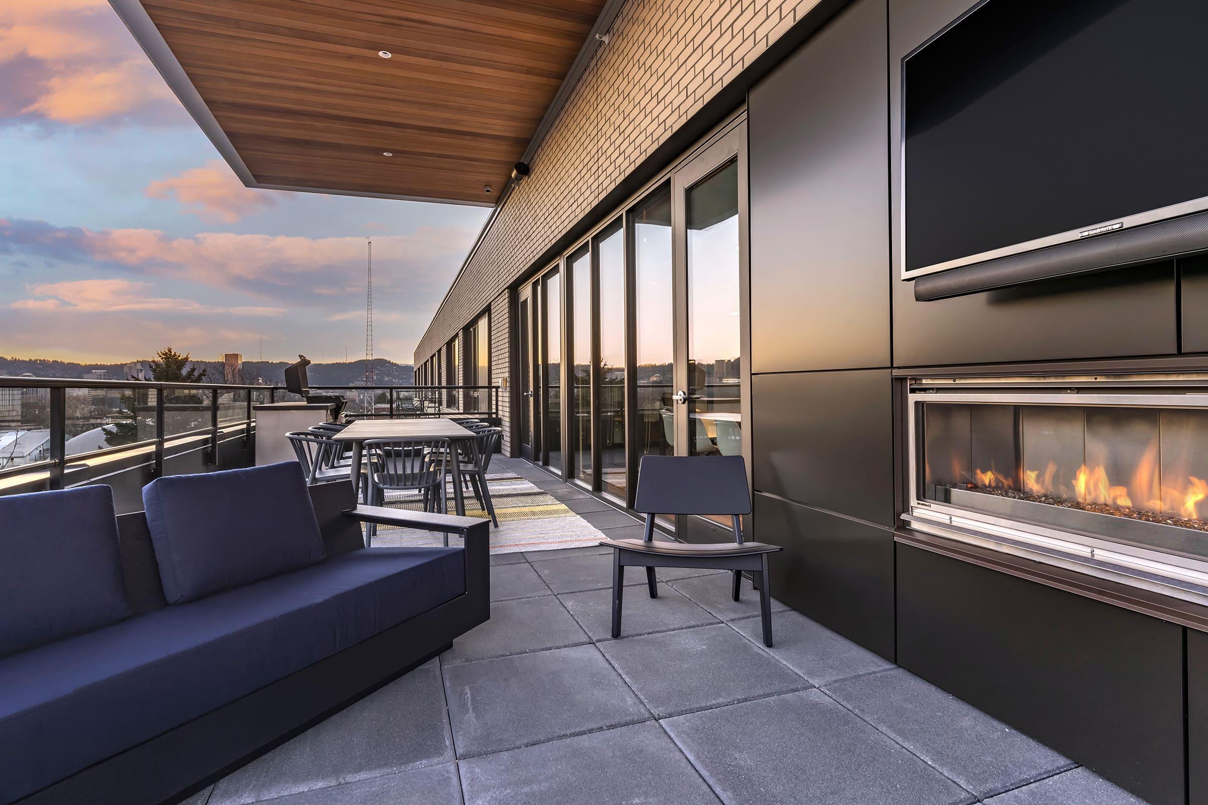 A modern rooftop terrace featuring a cozy seating area with a dark sofa and a chair, an outdoor fireplace, and a dining table. Large glass doors and windows provide views of the surroundings, while a warm sunset sky adds to the ambiance. The space is designed for relaxation and entertainment.