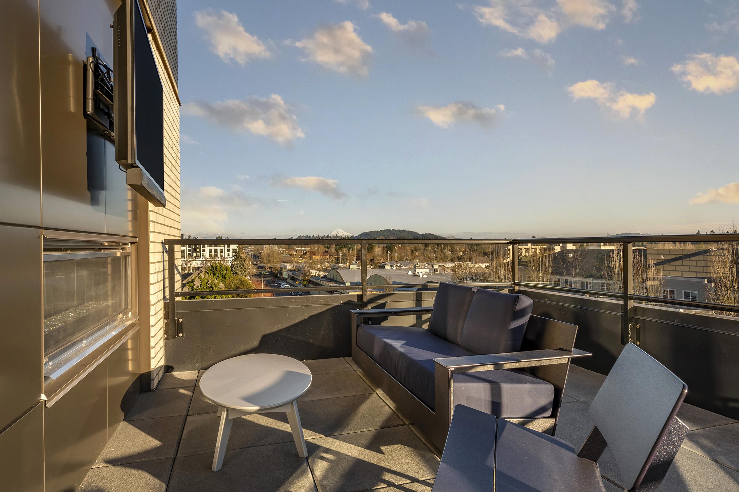 A modern rooftop patio featuring a comfortable blue sofa, a round white table, and a sleek chair. The backdrop includes a clear sky with a few clouds and distant mountains, offering a scenic view over nearby buildings and greenery.