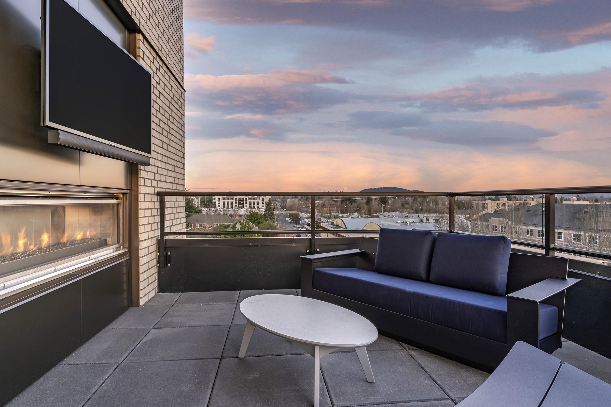 A modern balcony with a cozy outdoor seating area featuring a navy blue sofa, a round white coffee table, and a sleek outdoor fireplace. The backdrop captures a colorful sunset with clouds, enhancing the tranquil atmosphere of the urban setting.