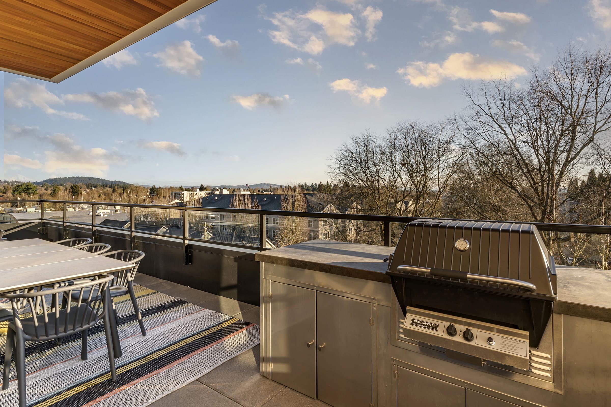 A modern outdoor patio featuring a barbecue grill, a dining table with chairs, and a rug. The space overlooks a scenic view of trees and buildings under a blue sky with clouds, suggesting a tranquil atmosphere for outdoor dining and relaxation.