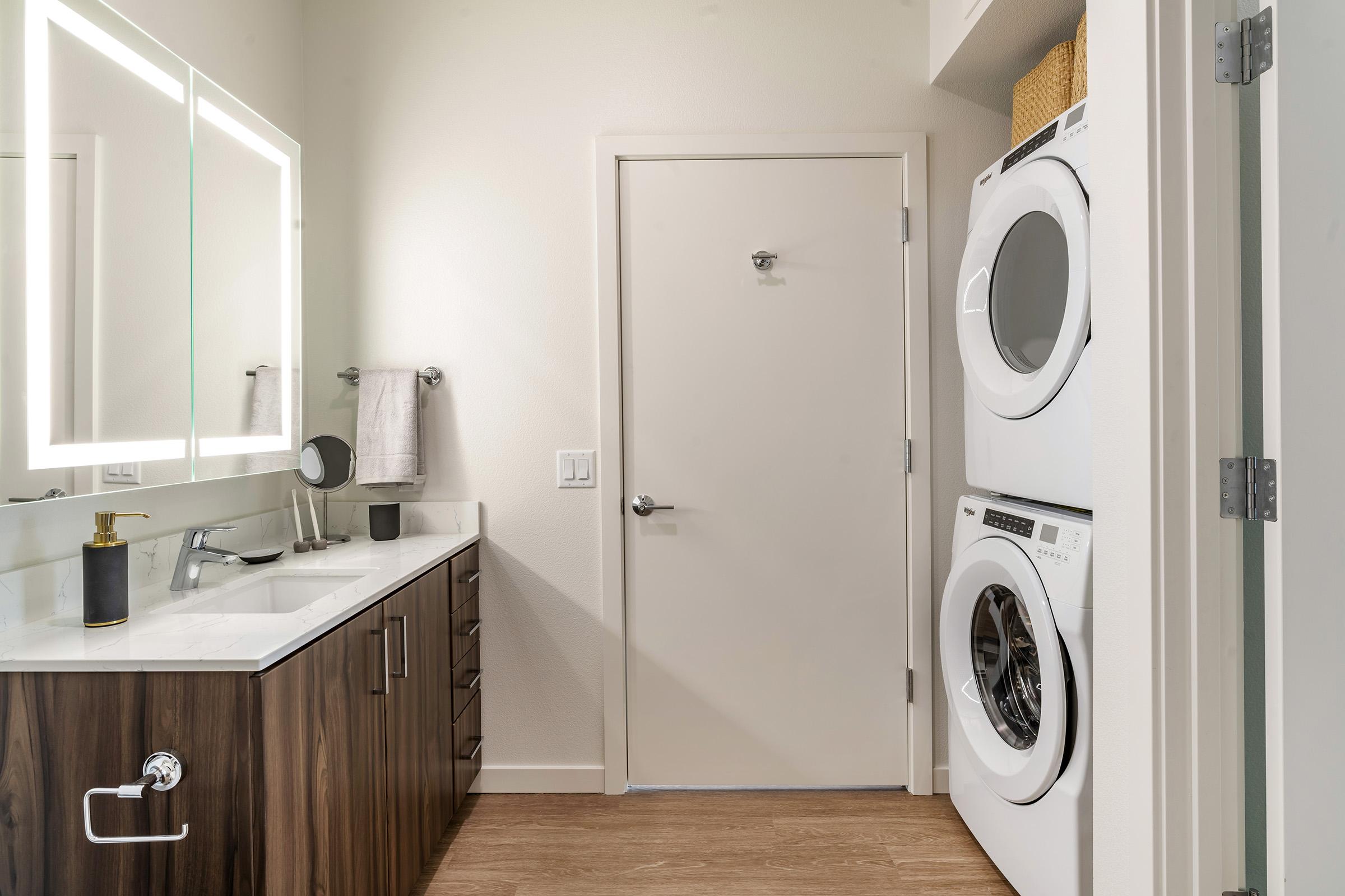 A modern laundry room with sleek cabinetry. It features a stackable washer and dryer on the right, a white door at the back, and a well-lit mirror above a vanity with a sink. A towel hangs on the wall, and there's a small round mirror on the countertop, creating a clean and organized look.