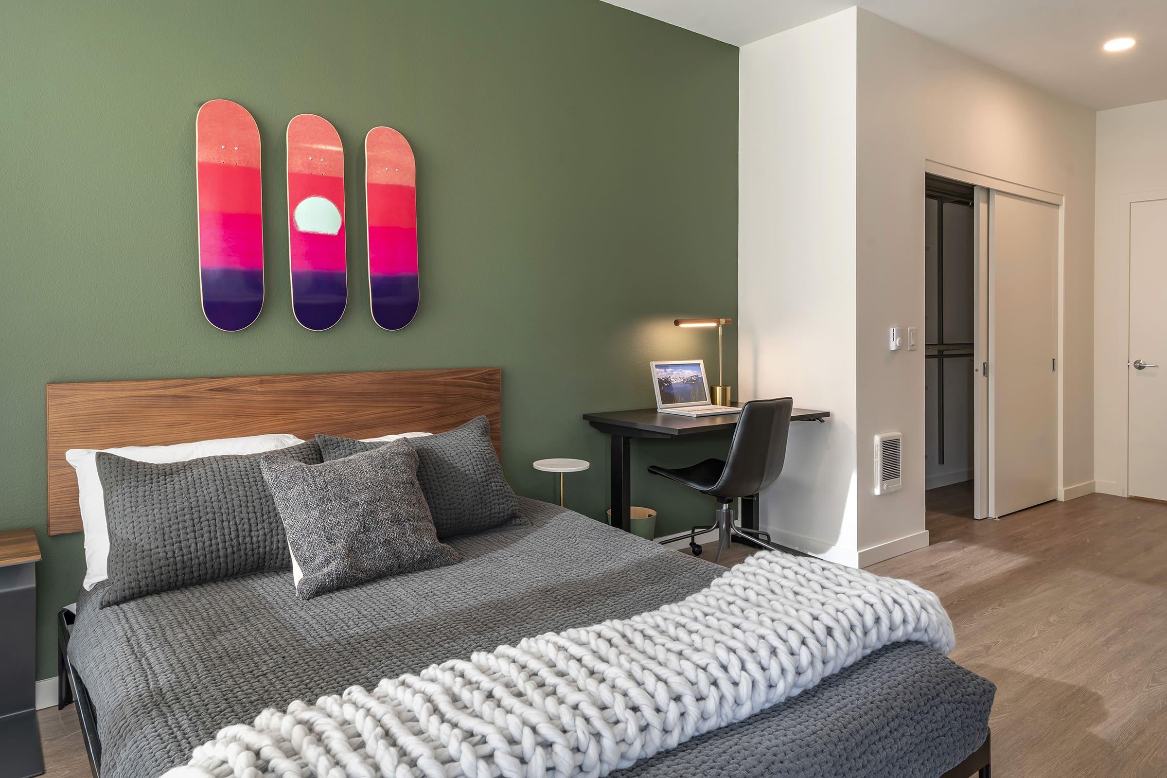 A cozy bedroom featuring a bed with gray bedding and decorative pillows. Above the bed, there are three colorful wall art pieces in the shape of skateboard decks, depicting a sunset. A small desk with a chair is positioned against a wall, and there's a doorway leading to another room. The walls are painted a calming green.