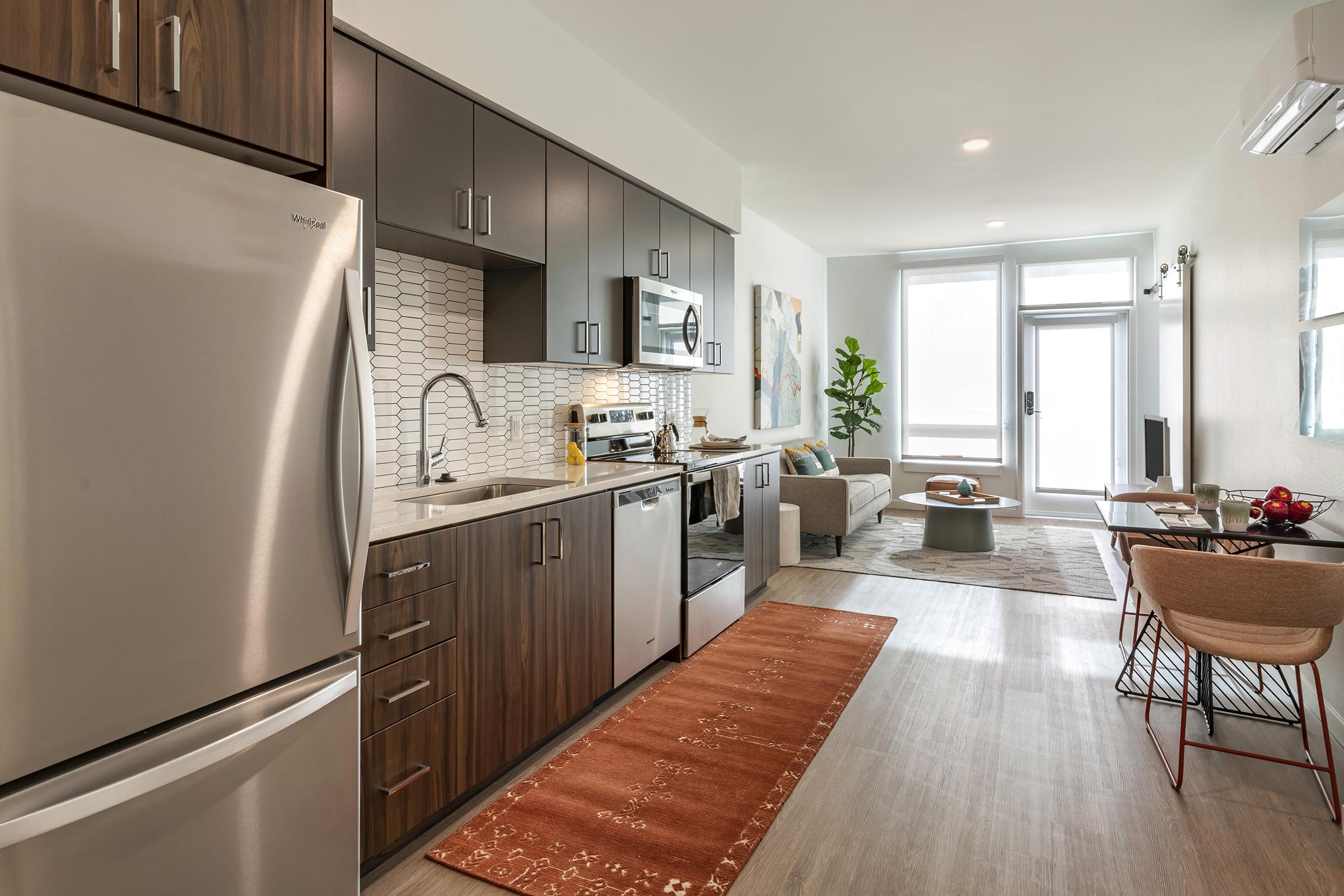 Modern kitchen and living area featuring stainless steel appliances, dark wooden cabinets, and a white tiled backsplash. A cozy sofa is positioned near a large window that allows natural light, with a coffee table and small dining area visible. Decorative plants and artwork add a stylish touch.