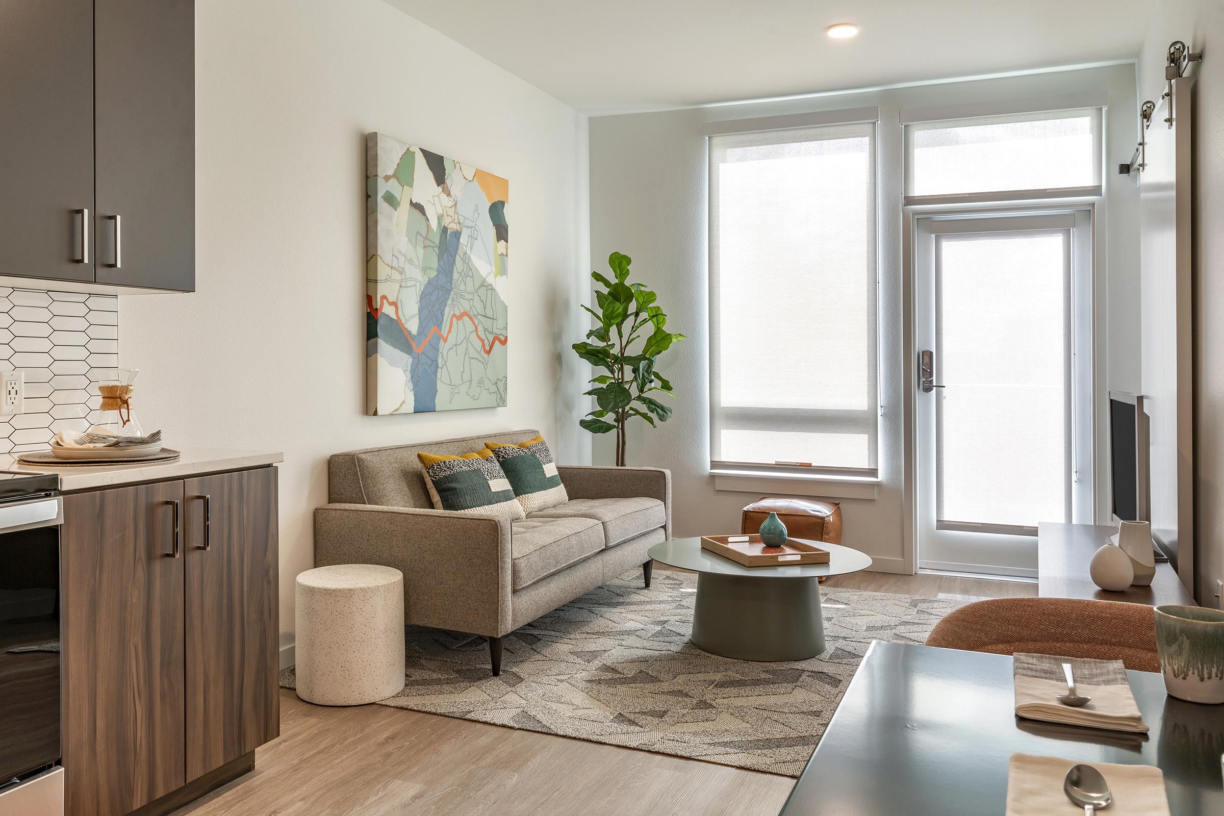 A modern living room featuring a light gray sofa with decorative pillows, a round coffee table, and a small plant. There's a large artwork on the wall, a sliding glass door leading outside, and stylish cabinetry with kitchen elements. The space has a cozy, inviting ambiance with natural light.