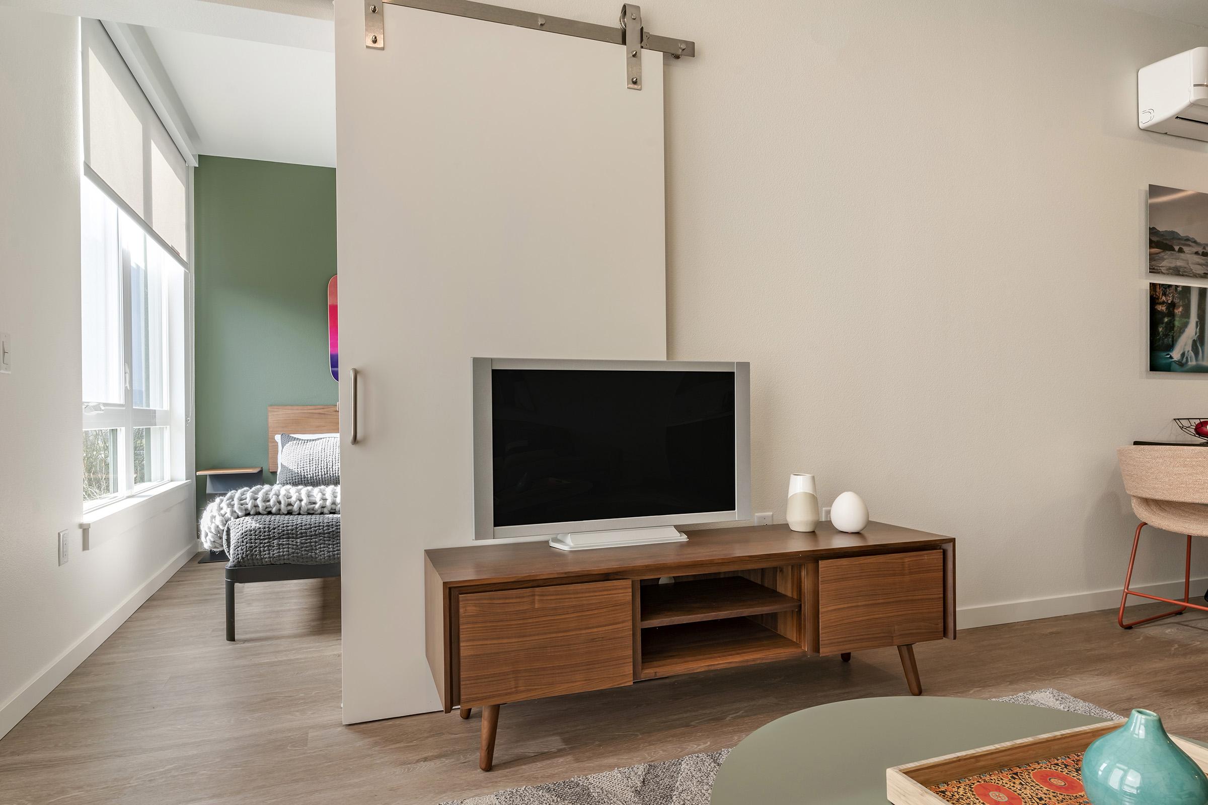 Modern living room featuring a wooden TV stand with a flat-screen television, decorative objects, and a beige sliding door that separates it from a bedroom. The room has neutral-colored walls and stylish furniture, providing a cozy and contemporary atmosphere.