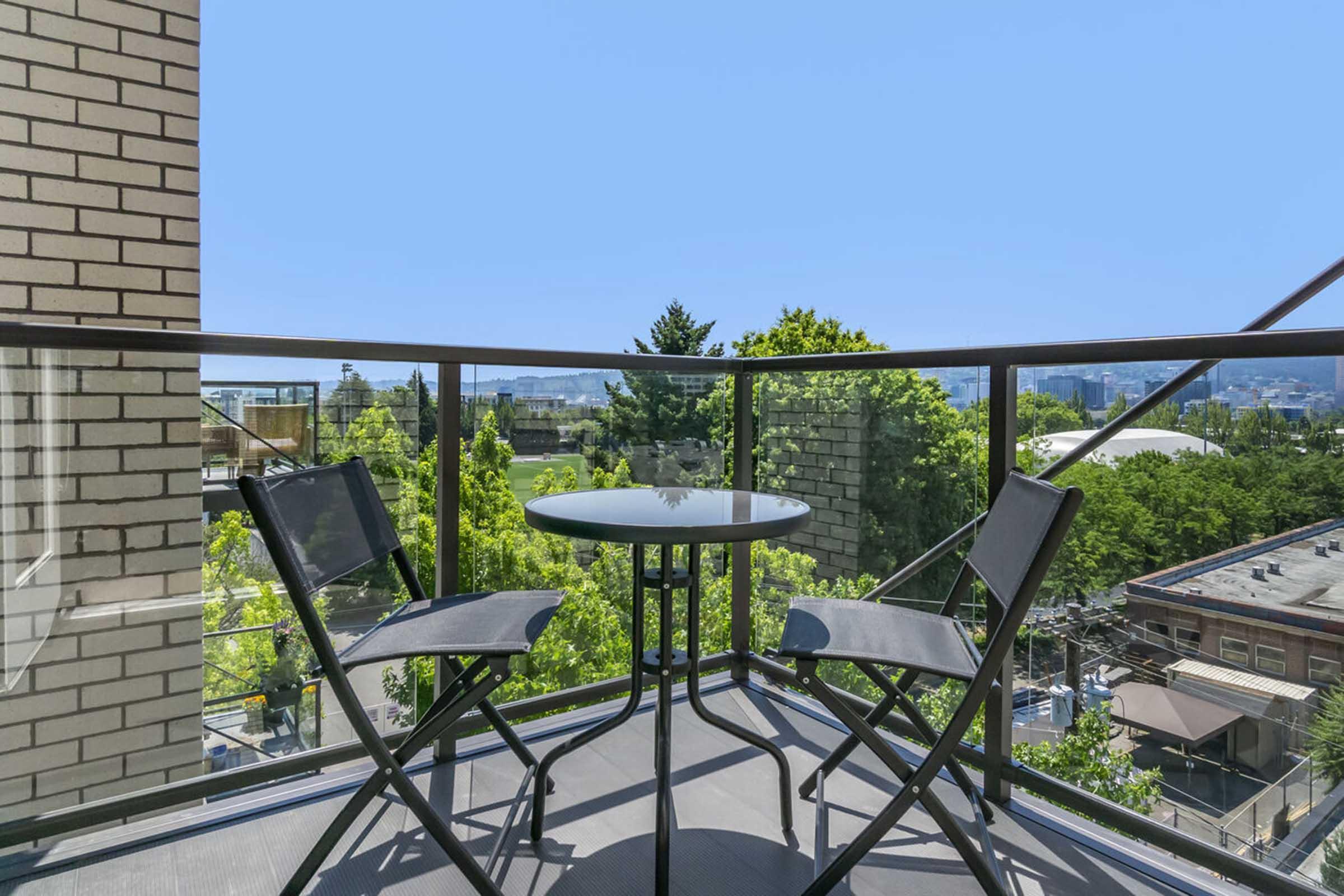 A small balcony featuring a round table and two folding chairs, surrounded by greenery. A clear blue sky is visible in the background, and the balcony overlooks a park-like area with trees and buildings in the distance. The glass railing offers an unobstructed view of the scenery.
