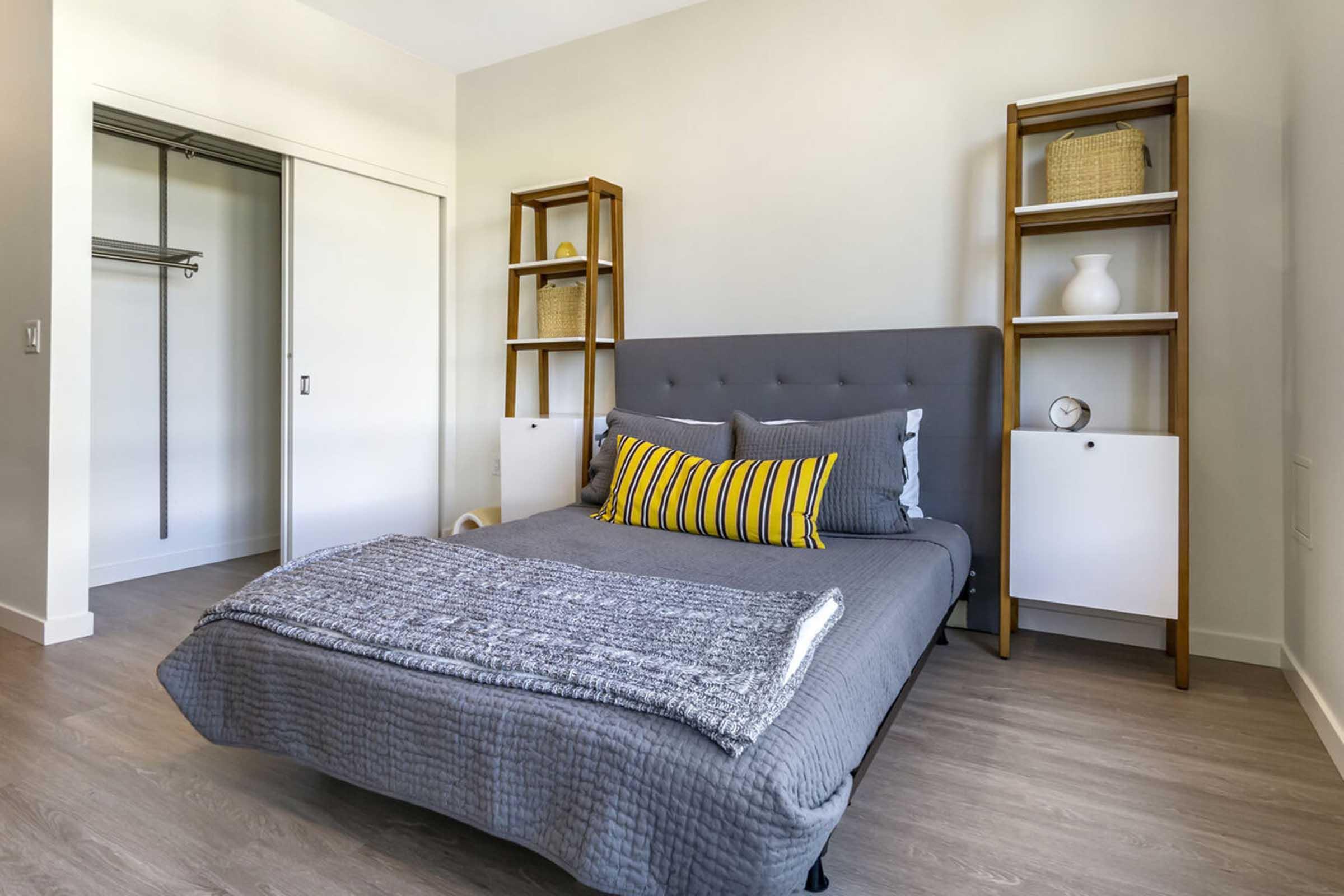 A modern bedroom featuring a gray bed with striped yellow and gray pillows, a textured throw blanket, and minimalistic wooden shelves. The room has light-colored walls, a closet with sliding doors, and simple decor, creating a clean and inviting atmosphere.
