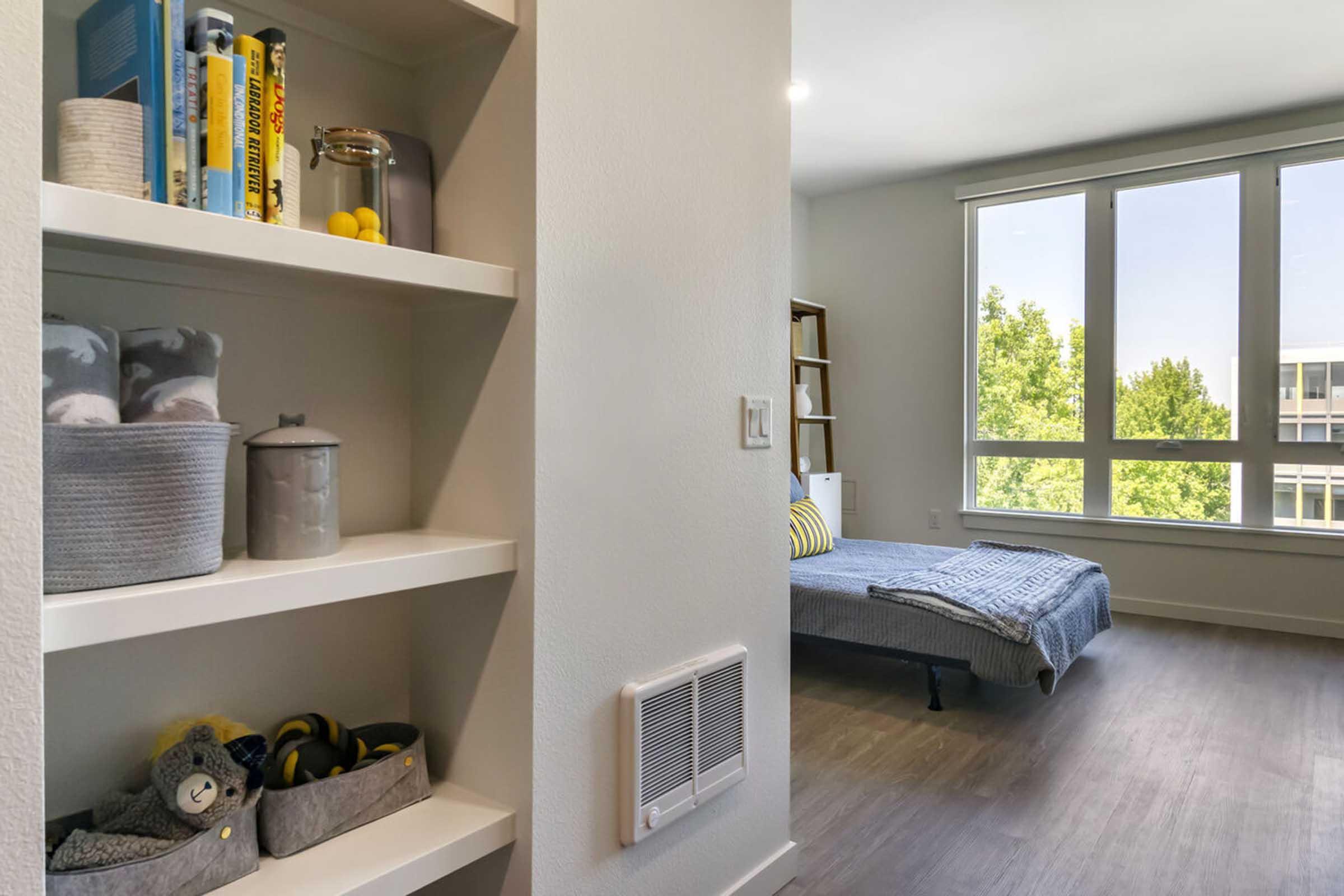 A view of a modern, well-lit room featuring a cozy bed with a blue blanket. To the left, a white shelf holds decorative items and storage baskets. Large windows allow natural light to fill the space, with greenery visible outside, creating a fresh and inviting atmosphere.