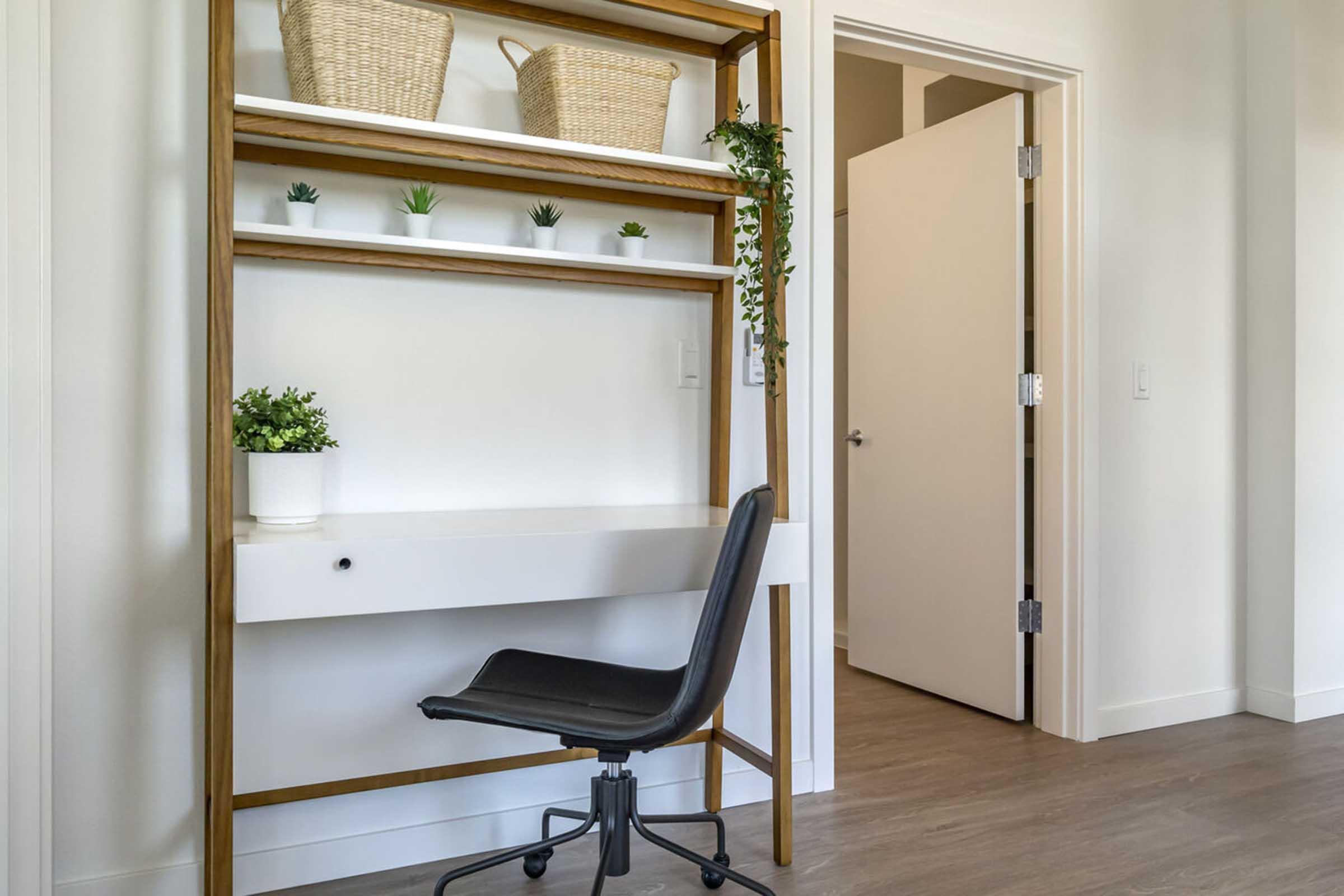 A minimalist workspace featuring a wooden shelf with two woven baskets and small potted plants. Below, a simple white desk contrasts with a black ergonomic chair. A doorway is visible in the background, leading to another room. The overall aesthetic is clean and modern, with neutral tones.