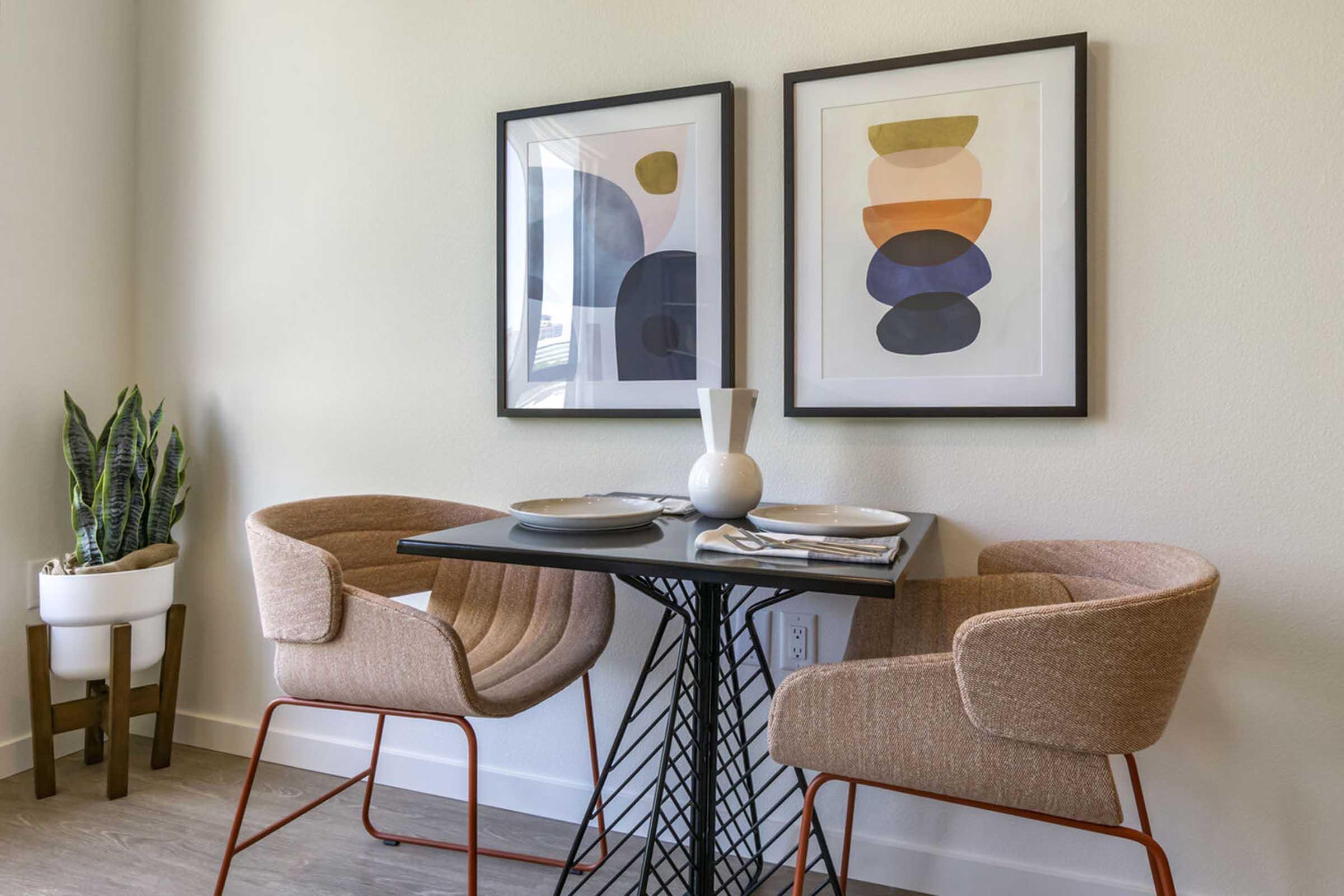 A cozy dining area featuring a round black table with two beige upholstered chairs. On the walls, there are two framed abstract art pieces with shapes in various neutral tones. A small green plant in a white pot is placed in the corner, enhancing the modern aesthetic of the space.