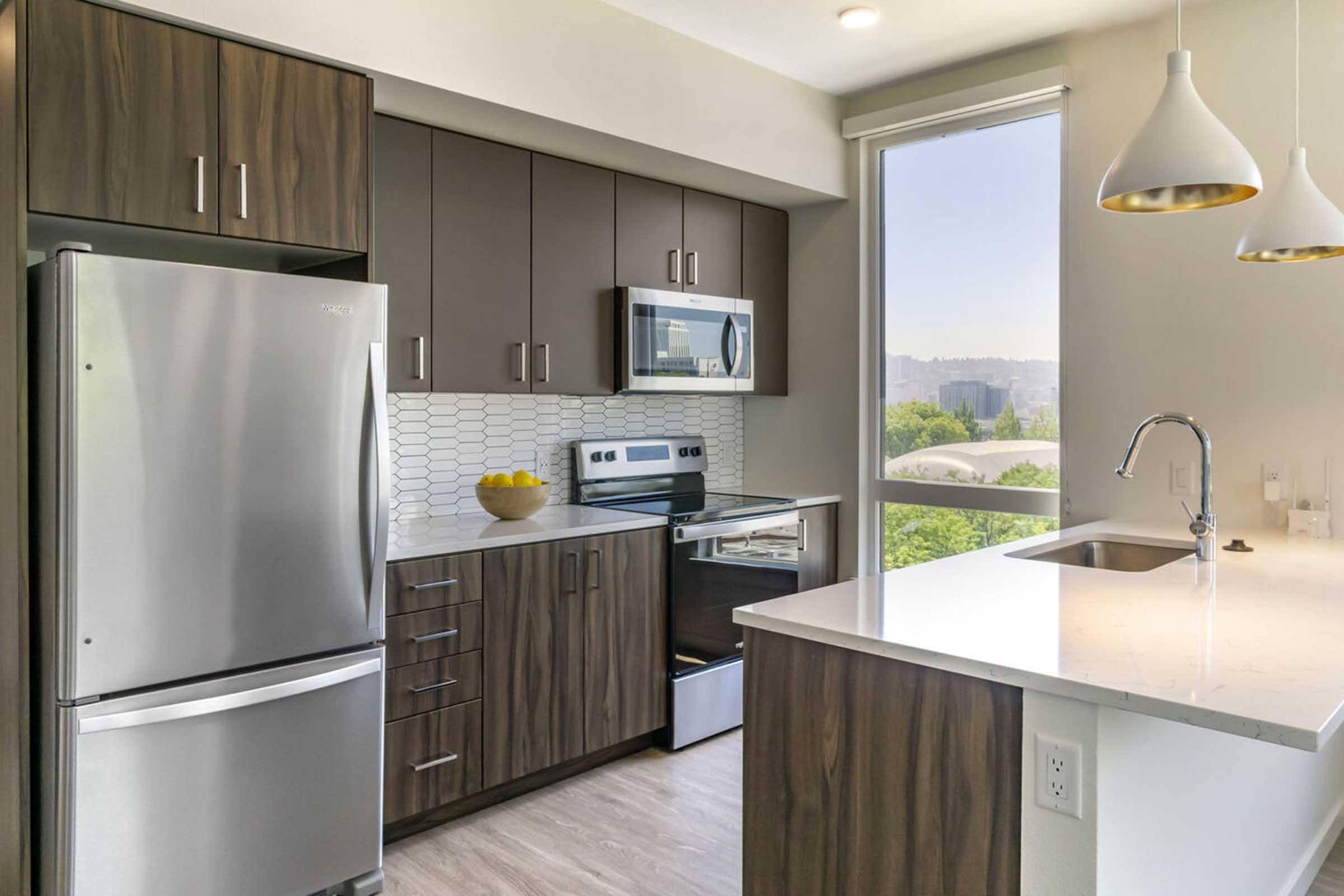 Modern kitchen featuring stainless steel appliances, including a refrigerator, oven, and microwave. Sleek dark wooden cabinetry complements a light countertop. A window offers a view of greenery outside, while pendant lights hang above the kitchen island.