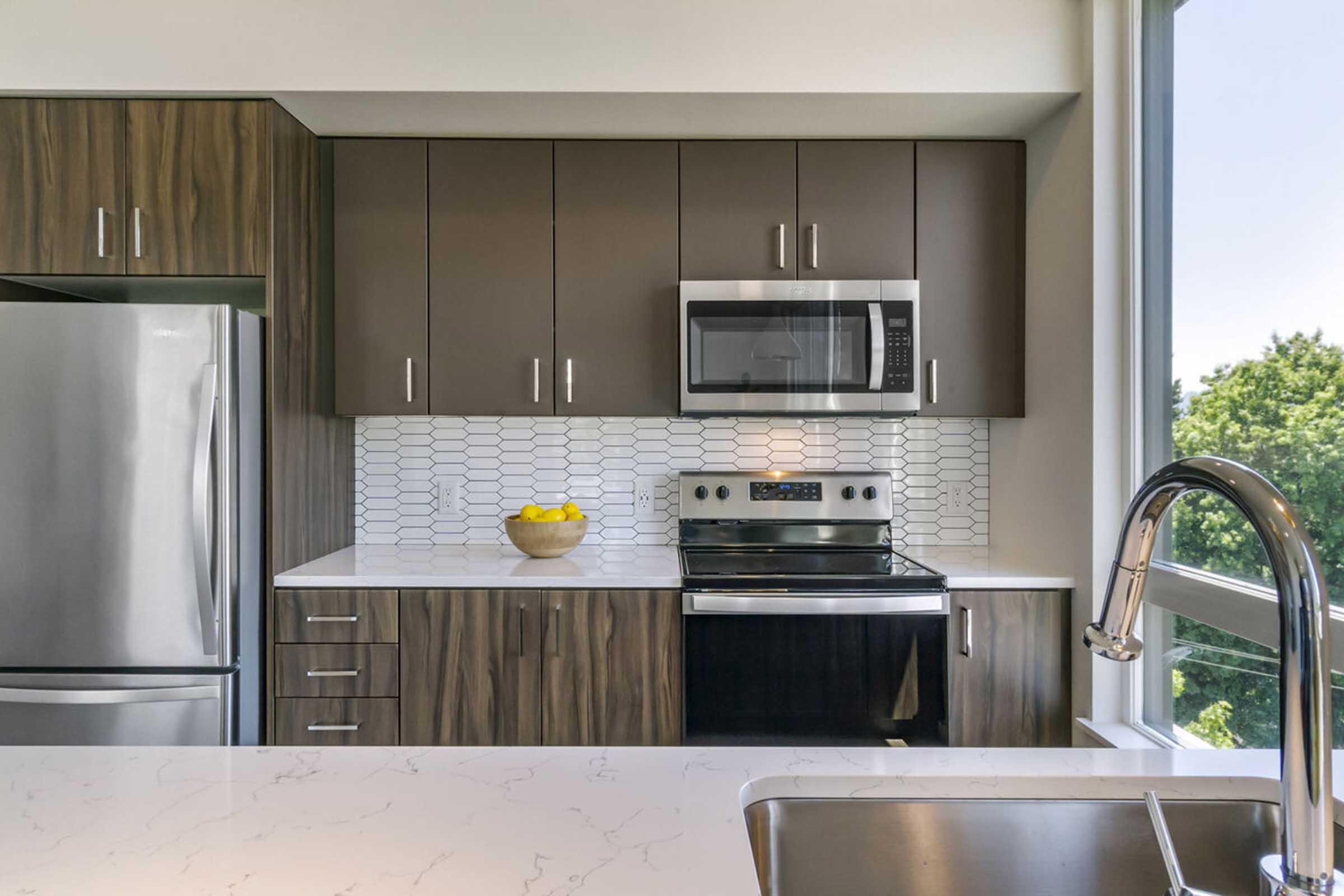 Modern kitchen featuring dark wood cabinetry, stainless steel appliances including a refrigerator, microwave, and oven. A white countertop and a white backsplash with a hexagonal pattern complement the design. A bowl of yellow lemons sits on the countertop near a stainless steel sink, with large windows providing natural light and a view of greenery outside.