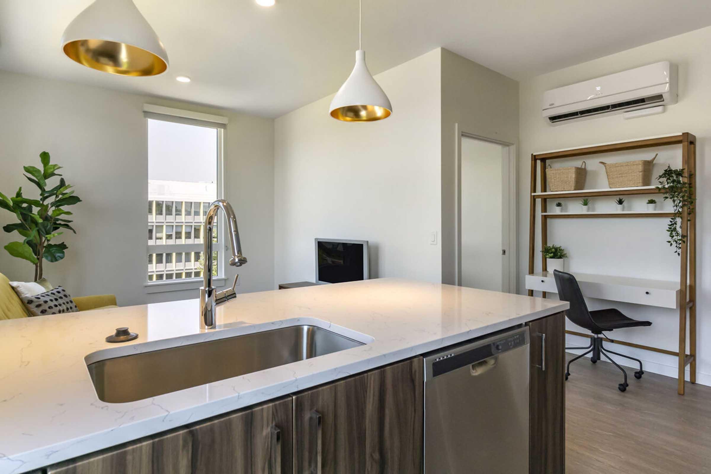 Modern kitchen with a sleek countertop and stainless steel dishwasher. A small living area features a green plant and a cozy sofa. There’s a workspace with a chair and shelves in the background. Two pendant lights hang from the ceiling, illuminating the open layout, and a window offers natural light.