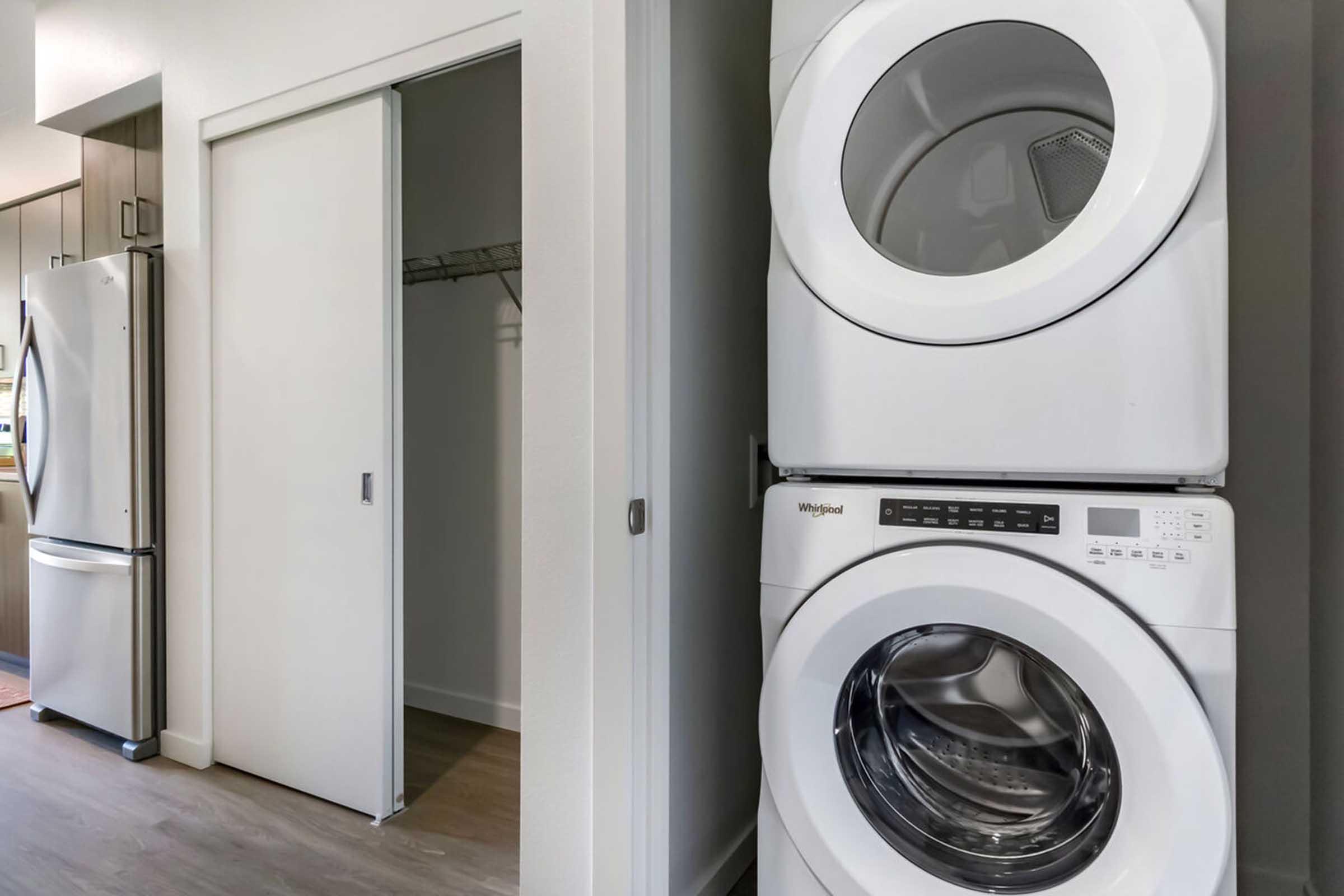 A compact laundry area featuring stacked white washer and dryer units. To the left, there's a modern refrigerator, and a partially open closet door reveals a hanger rod inside. The space is brightly lit with neutral-colored walls and flooring.