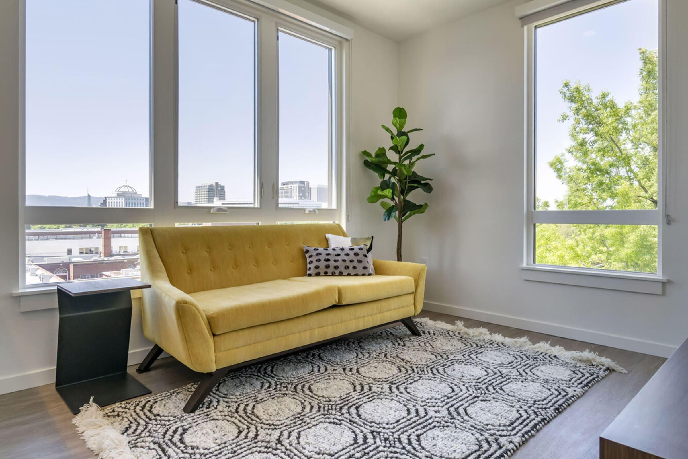 A modern living room featuring a vibrant yellow couch with a decorative pillow, a small table beside it, a leafy indoor plant, and large windows showing a cityscape. A textured area rug adds warmth to the space, which has a light, contemporary feel.