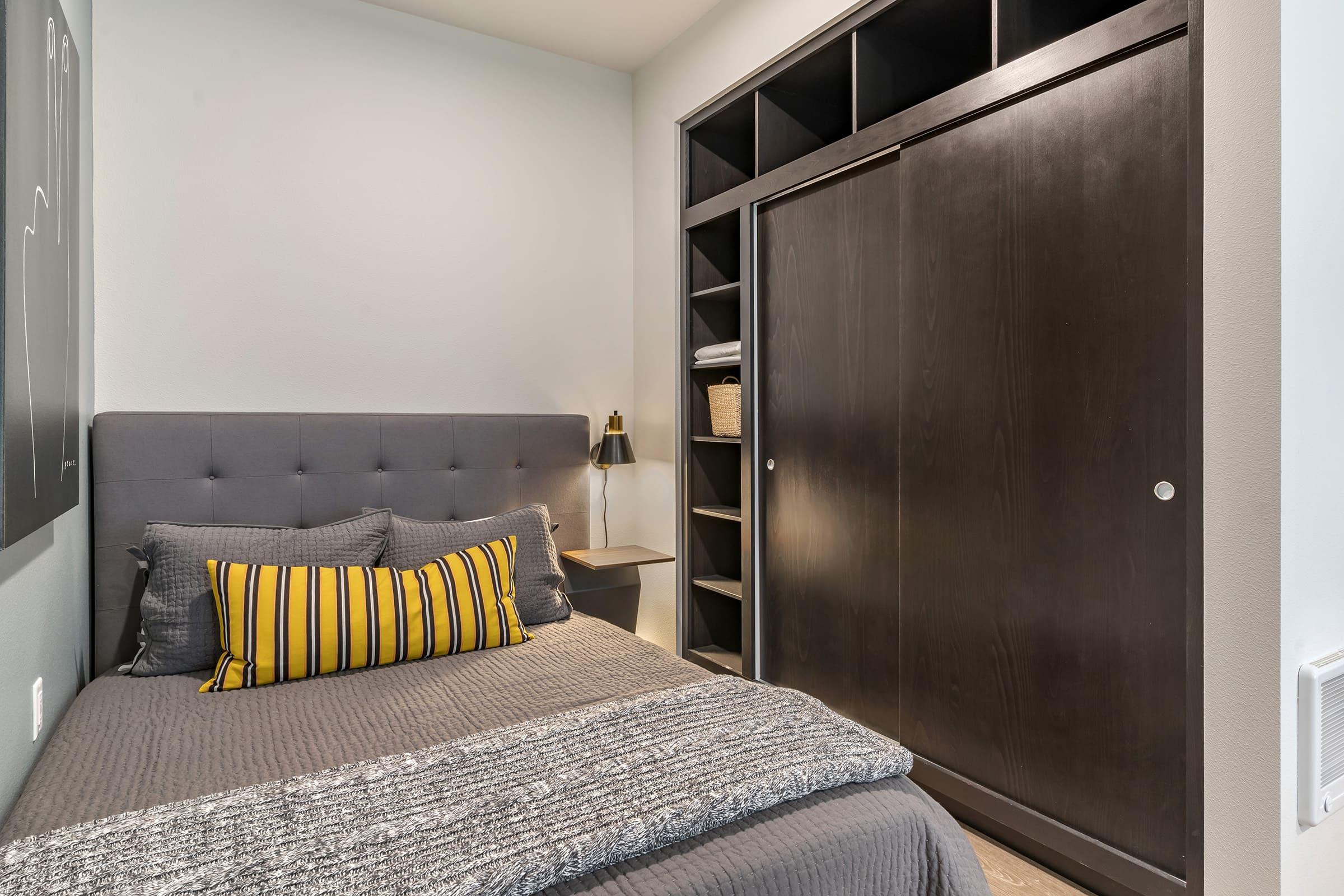 Cozy bedroom featuring a gray upholstered bed with striped yellow and gray pillows. A minimalist nightstand with a reading lamp is beside the bed, and a sleek dark wood closet with sliding doors occupies one wall. The room has neutral-colored walls and tidy shelves for storage.