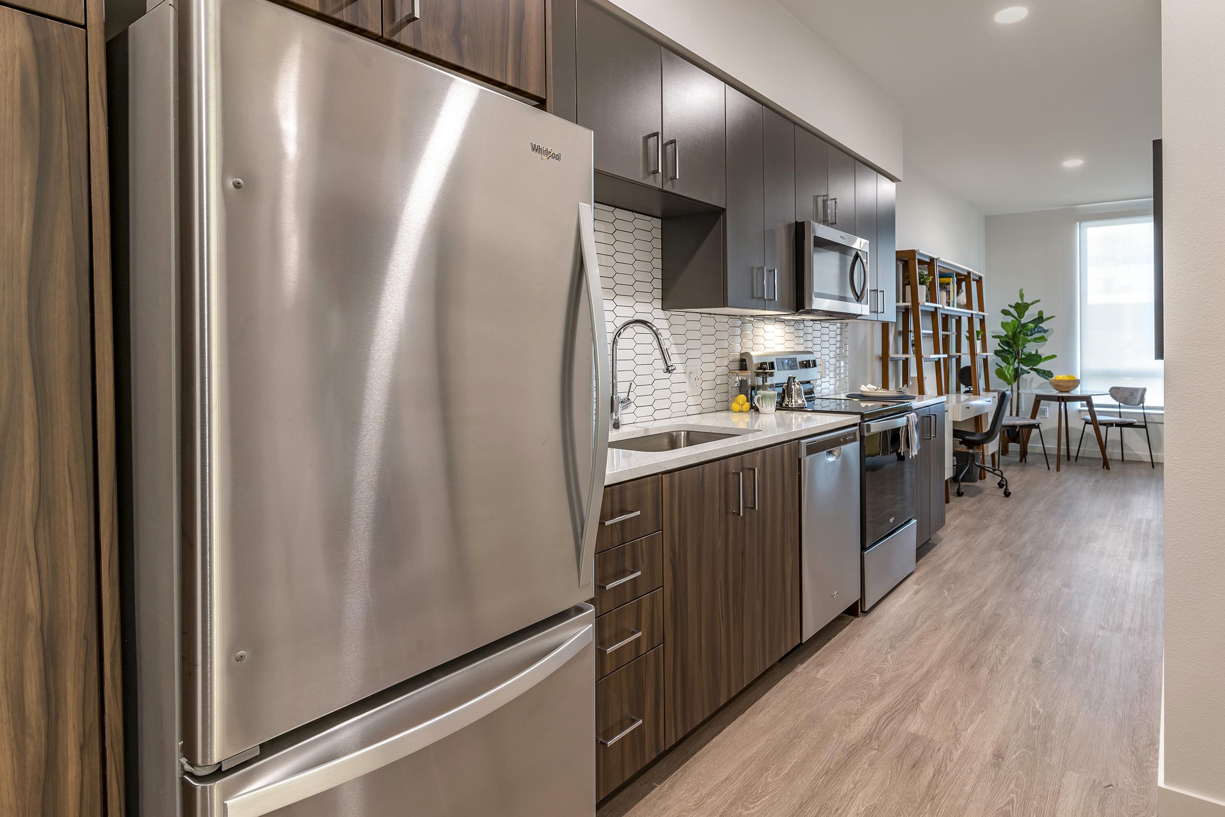 Modern kitchen featuring stainless steel appliances, including a refrigerator and microwave, with wooden cabinetry and a patterned backsplash. The space has a light, airy feel, complemented by a dining area with a table and chair set. Natural light flows in from a nearby window.