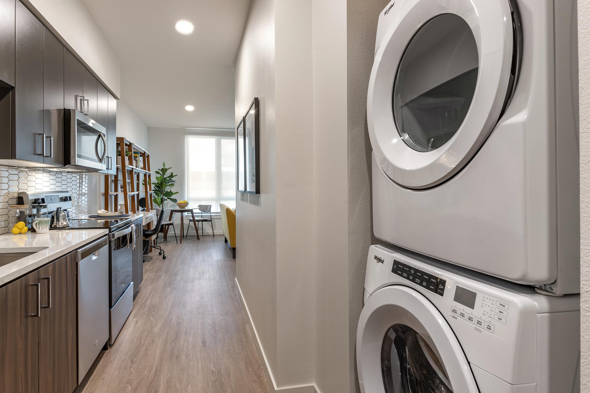 A modern laundry area with stacked white washer and dryer units, located in a stylish, open-concept apartment. The space includes dark wood cabinetry, a kitchen with stainless steel appliances, and a glimpse of a cozy workspace and plant in the background.