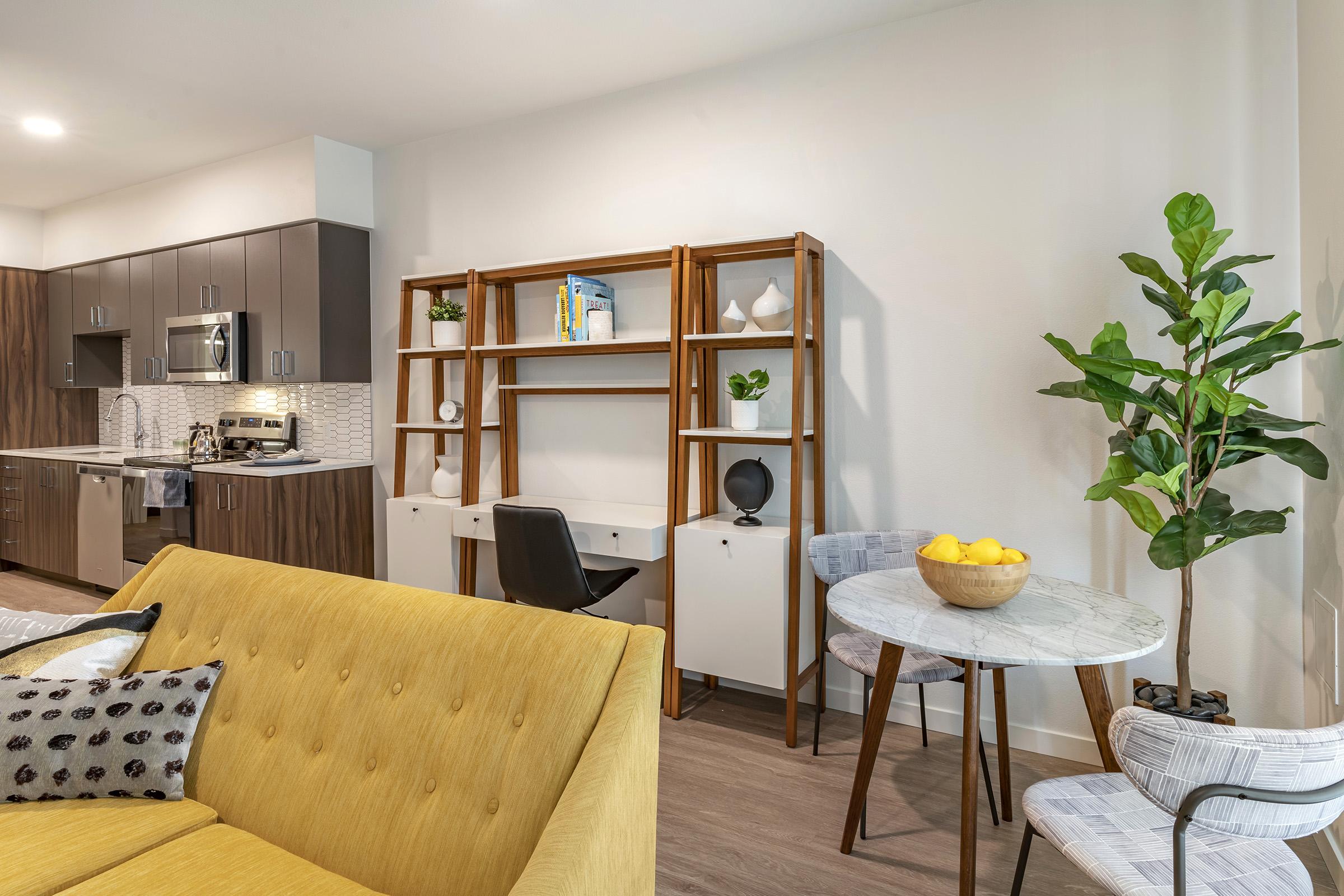 Modern interior of a living space featuring a yellow couch, a wooden shelf with decorative items, a small round table with a marble top and a bowl of lemons, and a potted plant. The kitchen area is visible in the background with dark cabinetry and stainless steel appliances.