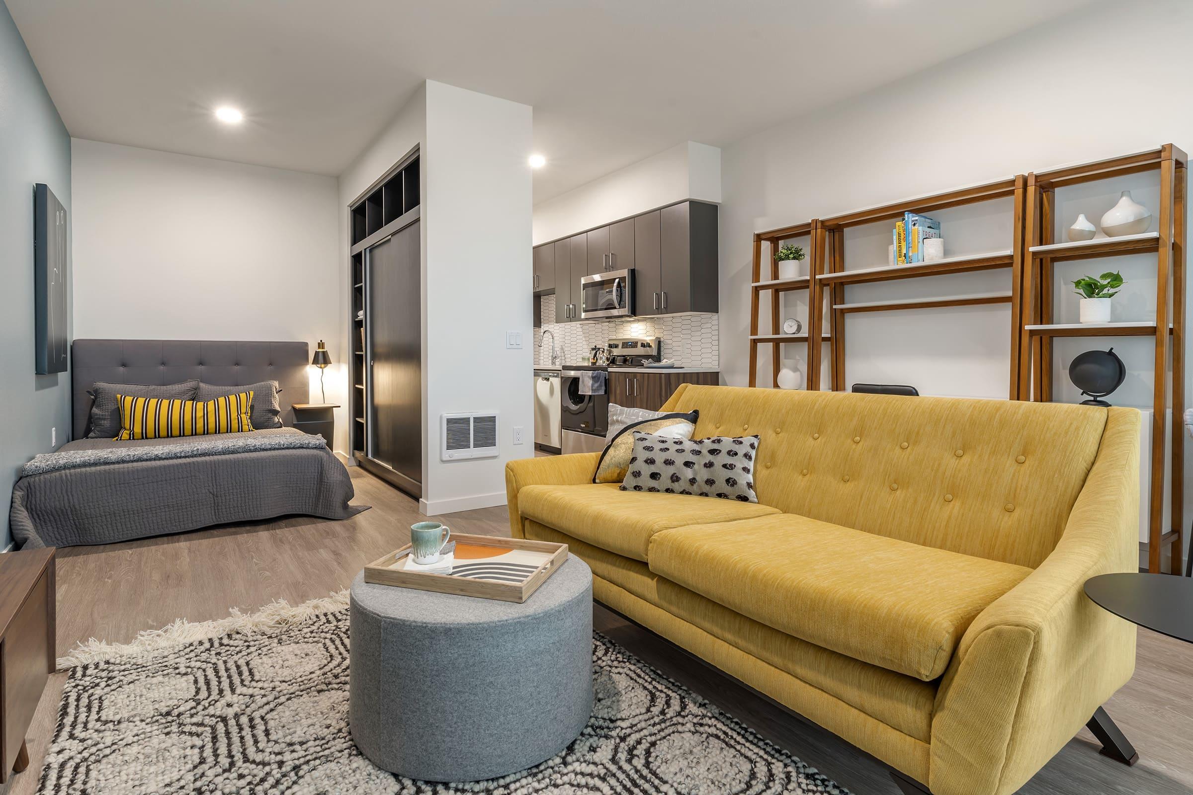 A modern living space featuring a yellow sofa, gray armchair, and a round coffee table on a patterned rug. In the background, there's a gray bed with decorative pillows and a stylish kitchen area with black and silver appliances. Bookshelves display décor items and plants, creating a cozy atmosphere.