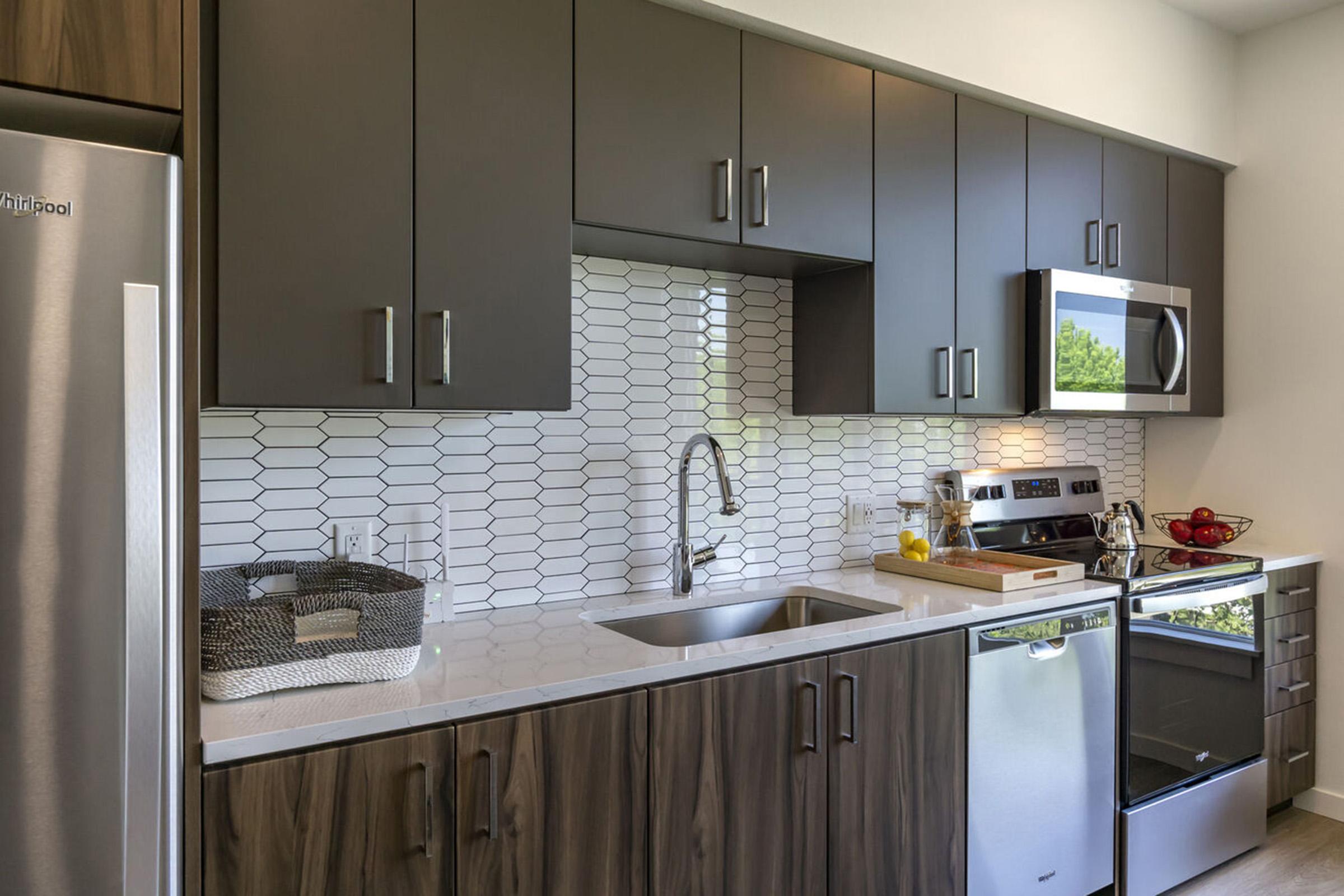 Modern kitchen featuring sleek dark cabinetry, a stainless steel refrigerator, a built-in microwave, and an electric stove. The countertop is made of light-colored stone, with a stylish backsplash of hexagonal tiles. A sink is installed beneath the cabinets, with minimal decorative items on the countertop.
