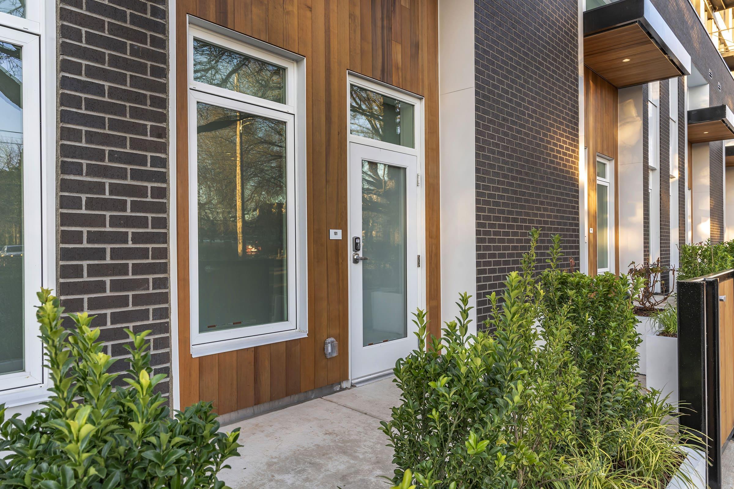 Modern building entrance featuring a white door surrounded by large windows, set against a backdrop of warm wooden paneling and dark brick. The entrance is flanked by neatly trimmed green shrubs and plants, creating a welcoming aesthetic.