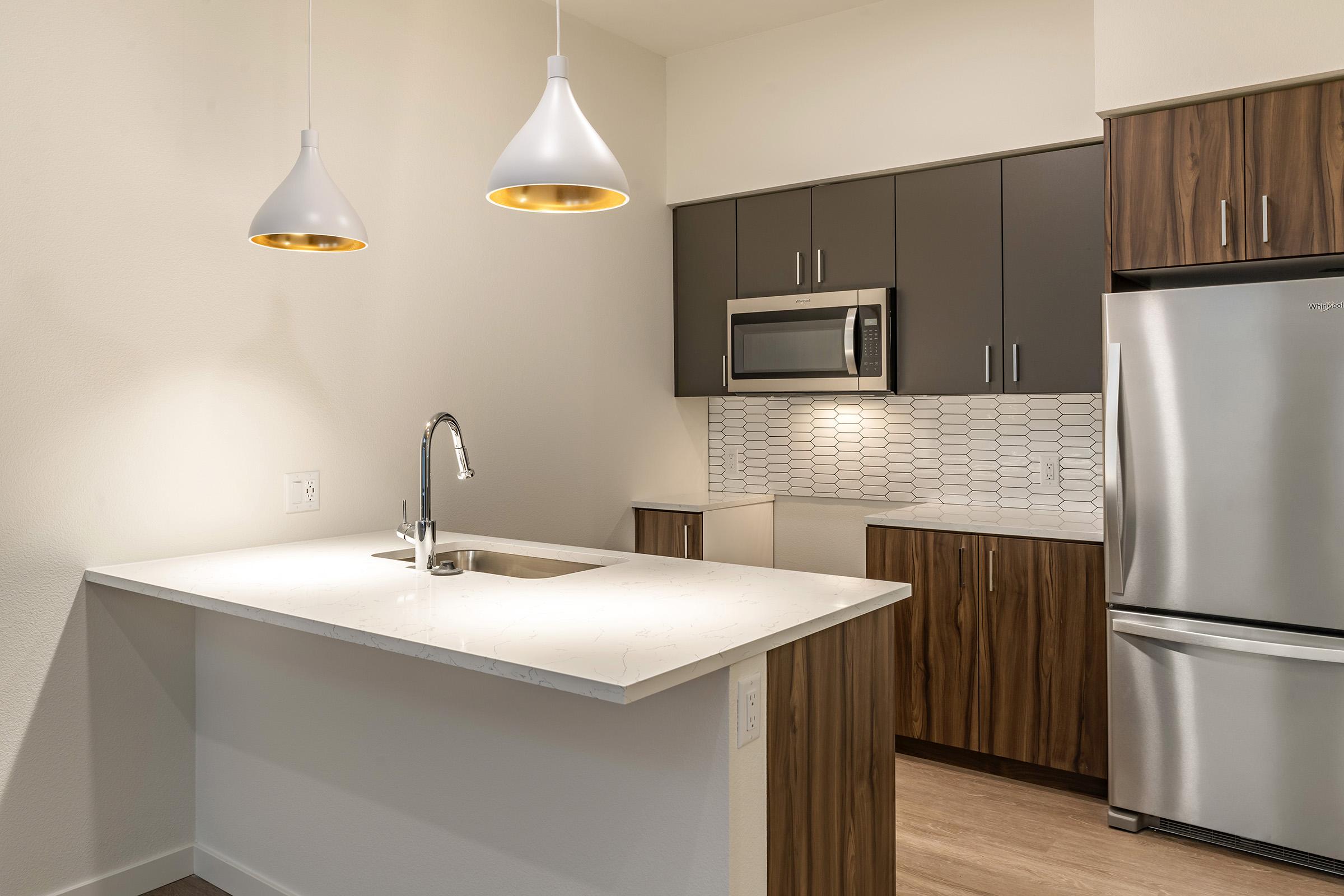 Modern kitchen featuring a light-colored countertop with a sink, dark cabinetry, a stainless steel refrigerator, and a microwave. Two pendant lights hang above the countertop, and a patterned backsplash complements the sleek design. The flooring is wood, creating a warm atmosphere.