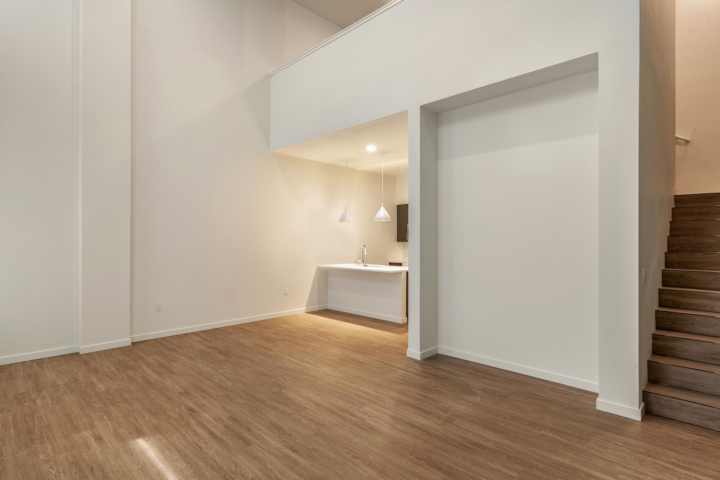 A spacious, empty room with light-colored walls and hardwood flooring. On one side, there is a small kitchen area with pendant lighting and a countertop. A staircase leads to an upper level. The room is bright, with natural light coming from above.
