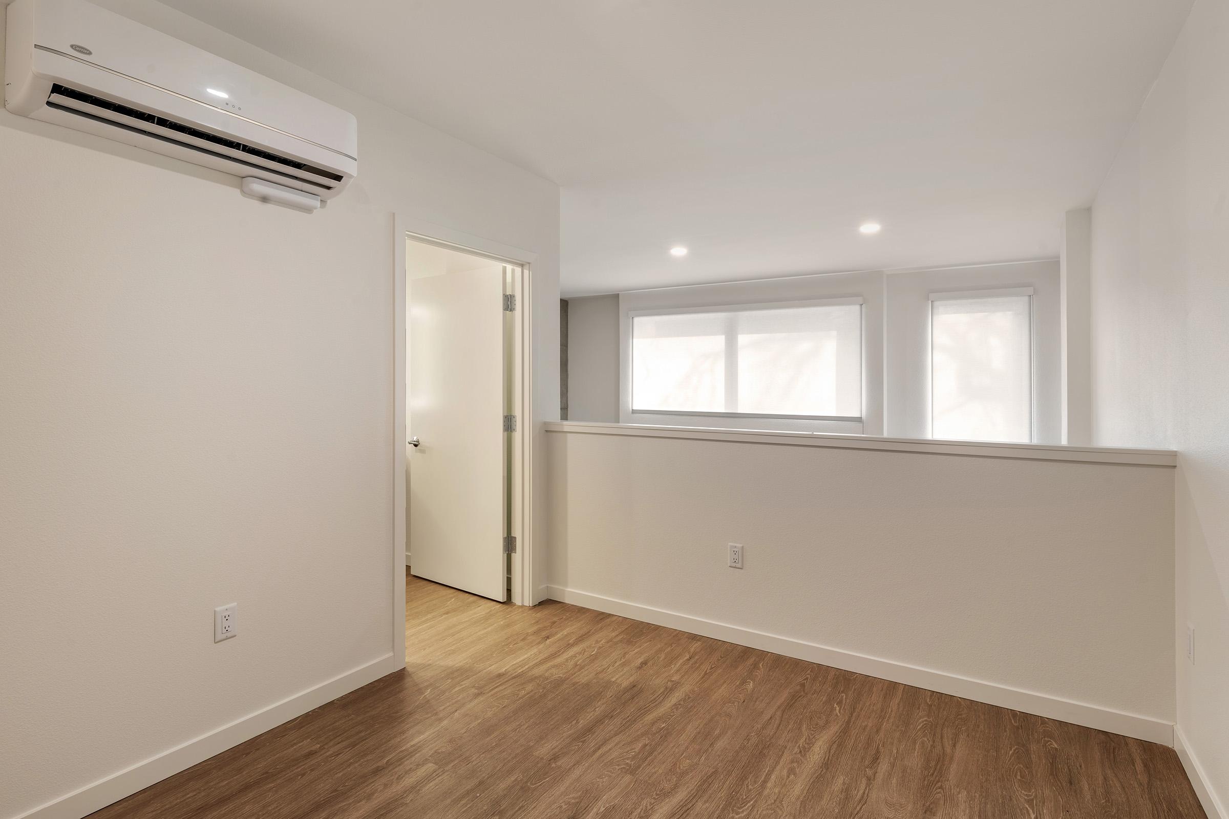 Empty interior space with light wood flooring. There is an air conditioning unit mounted on the wall and a door leading to another room. Large windows allow natural light to fill the area, creating a bright and open atmosphere. The walls are painted in a neutral color, enhancing the sense of openness.
