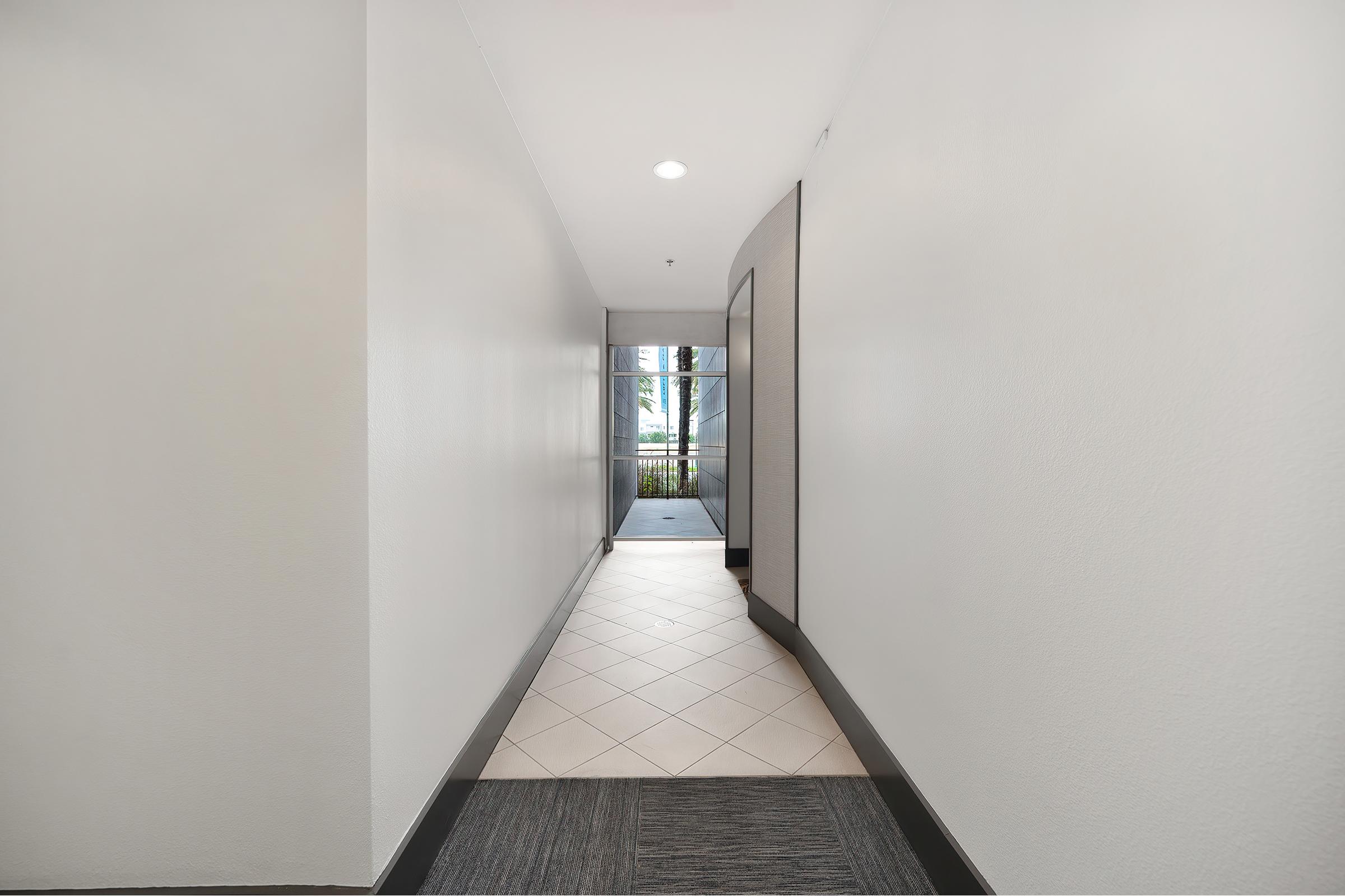 A well-lit hallway with white walls and a tiled floor, leading to an entrance with large windows that provide a view of the outdoors. The hallway has a modern design, featuring a mix of tile and carpet near the entrance.