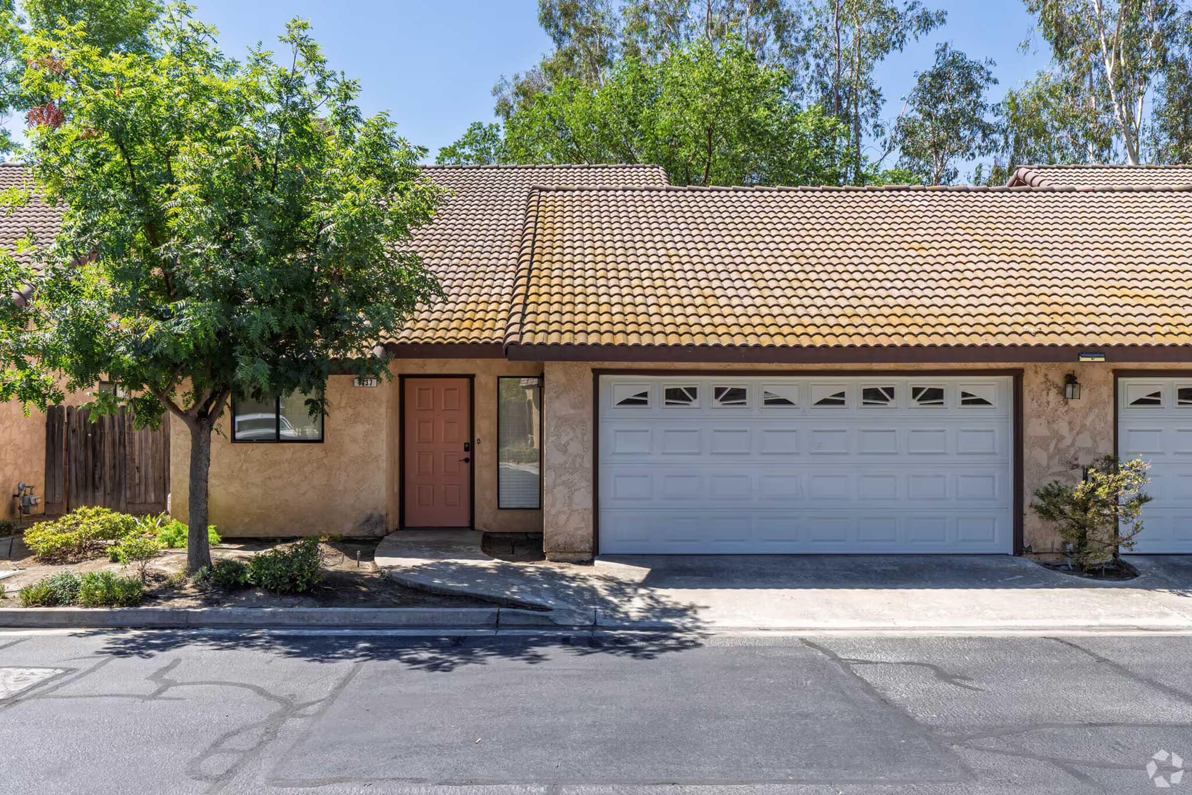 A residential building with a brown tile roof, featuring a light pink front door and a two-car garage. The surrounding area includes a small landscaped garden with trees and shrubs, set against a clear blue sky. The driveway is paved, leading to the garage area.