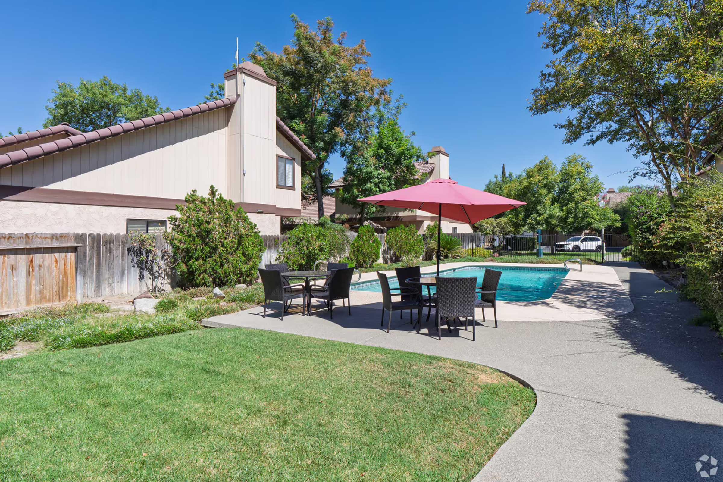 A sunny backyard with a swimming pool surrounded by a concrete patio. There's a dining table set with black chairs and a large red umbrella for shade. Lush green grass and shrubbery enhance the outdoor space, while two residential buildings are visible in the background against a clear blue sky.