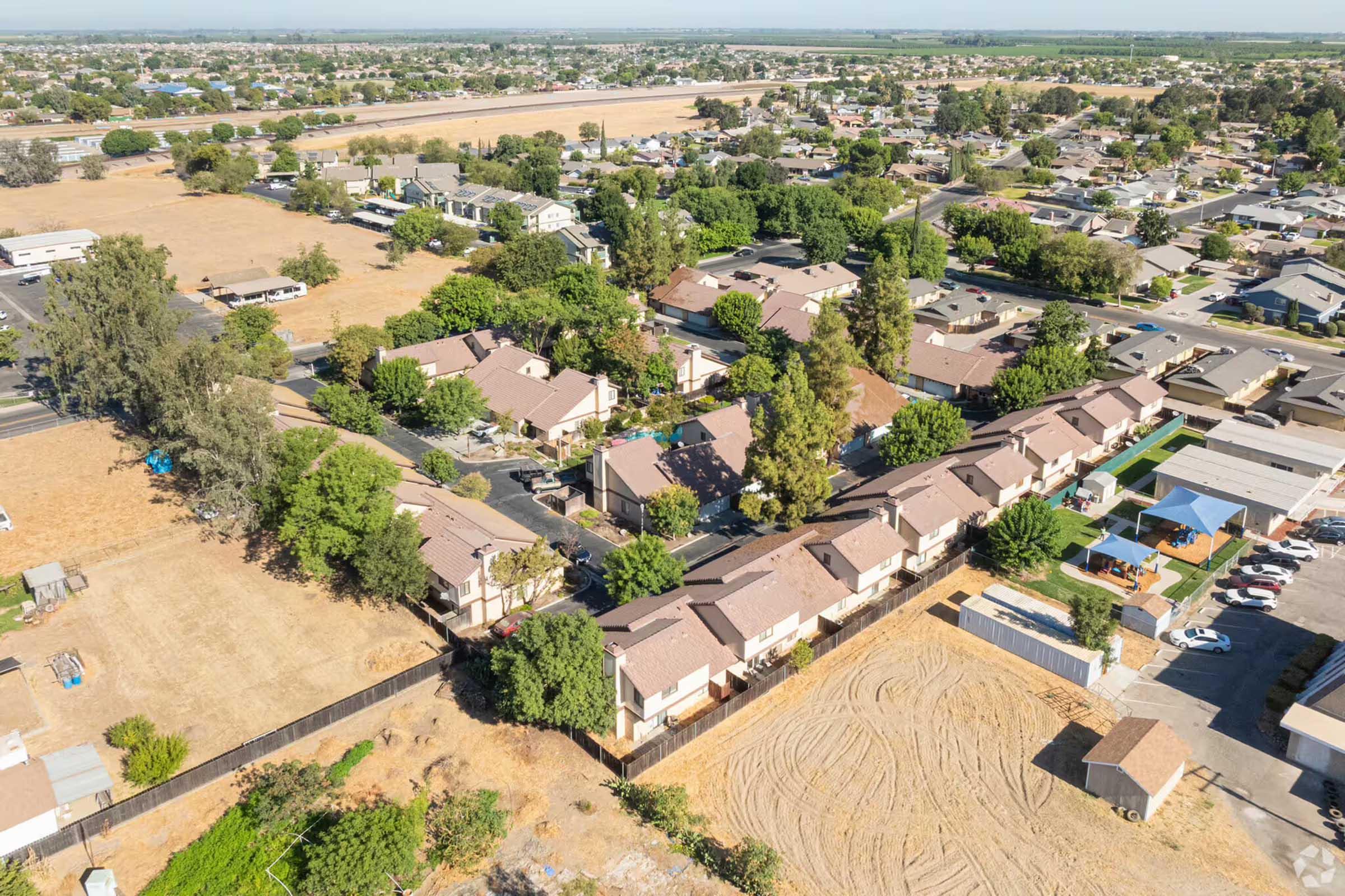 Aerial view of a neighborhood featuring residential buildings and green trees. There are paved roads and some areas of dry grass. In the background, more houses can be seen along with a clear blue sky. The layout shows a mix of housing styles and open spaces, typical of suburban development.