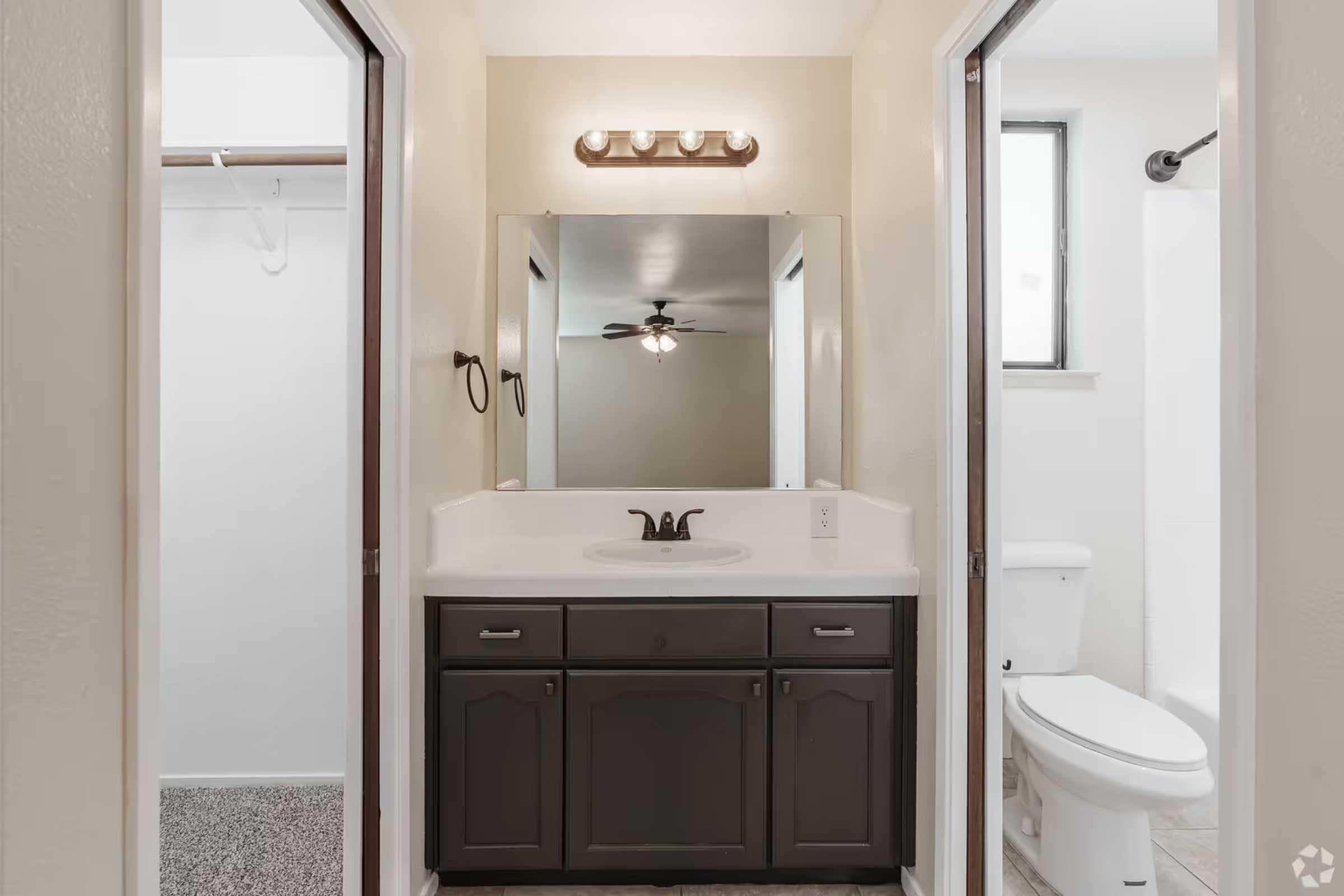 A well-lit bathroom featuring a double-sink vanity with a large mirror above. On one side, there's a white toilet and a shower area with a glass door. The other side shows a closet with a hanging rod. The walls are painted a soft beige, and there is a ceiling fan overhead.