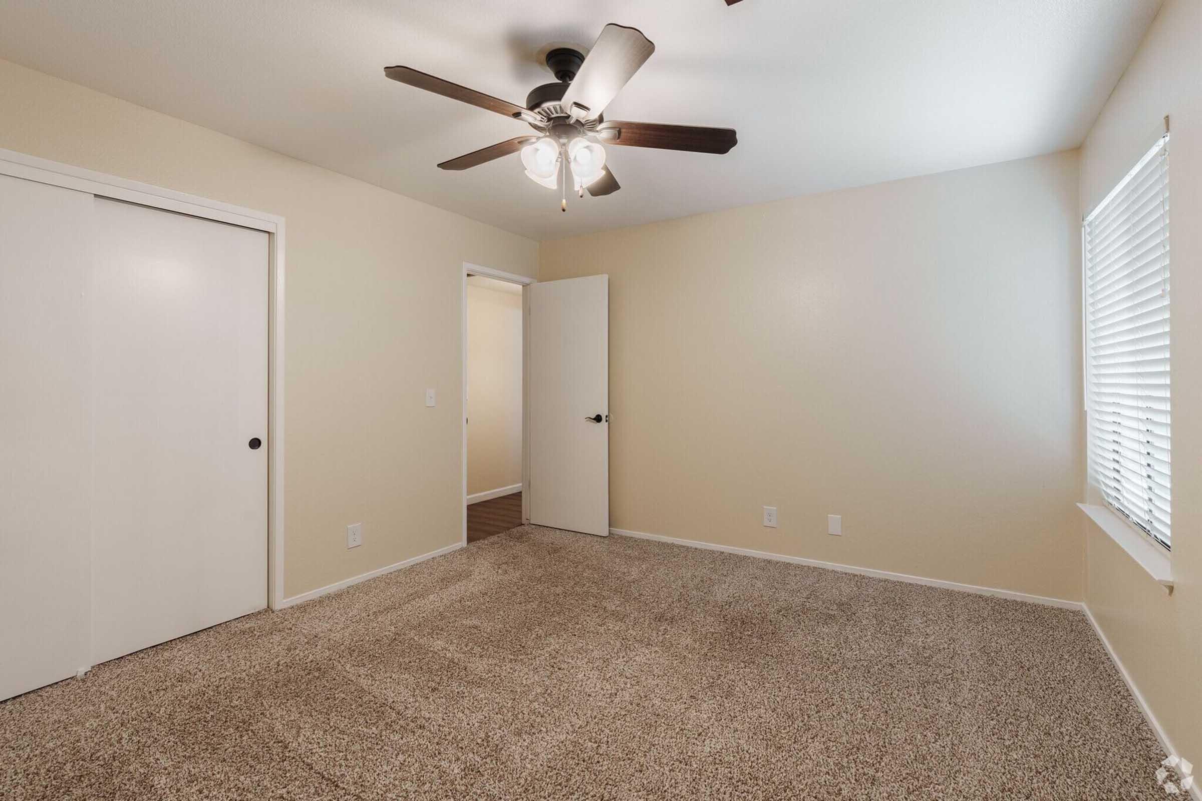 Empty room featuring beige walls and carpet flooring. A ceiling fan with light hangs from the ceiling. A door leads to another room, while a window with blinds provides natural light. There are two closed closet doors on one wall. Overall, the room has a clean and simple appearance.