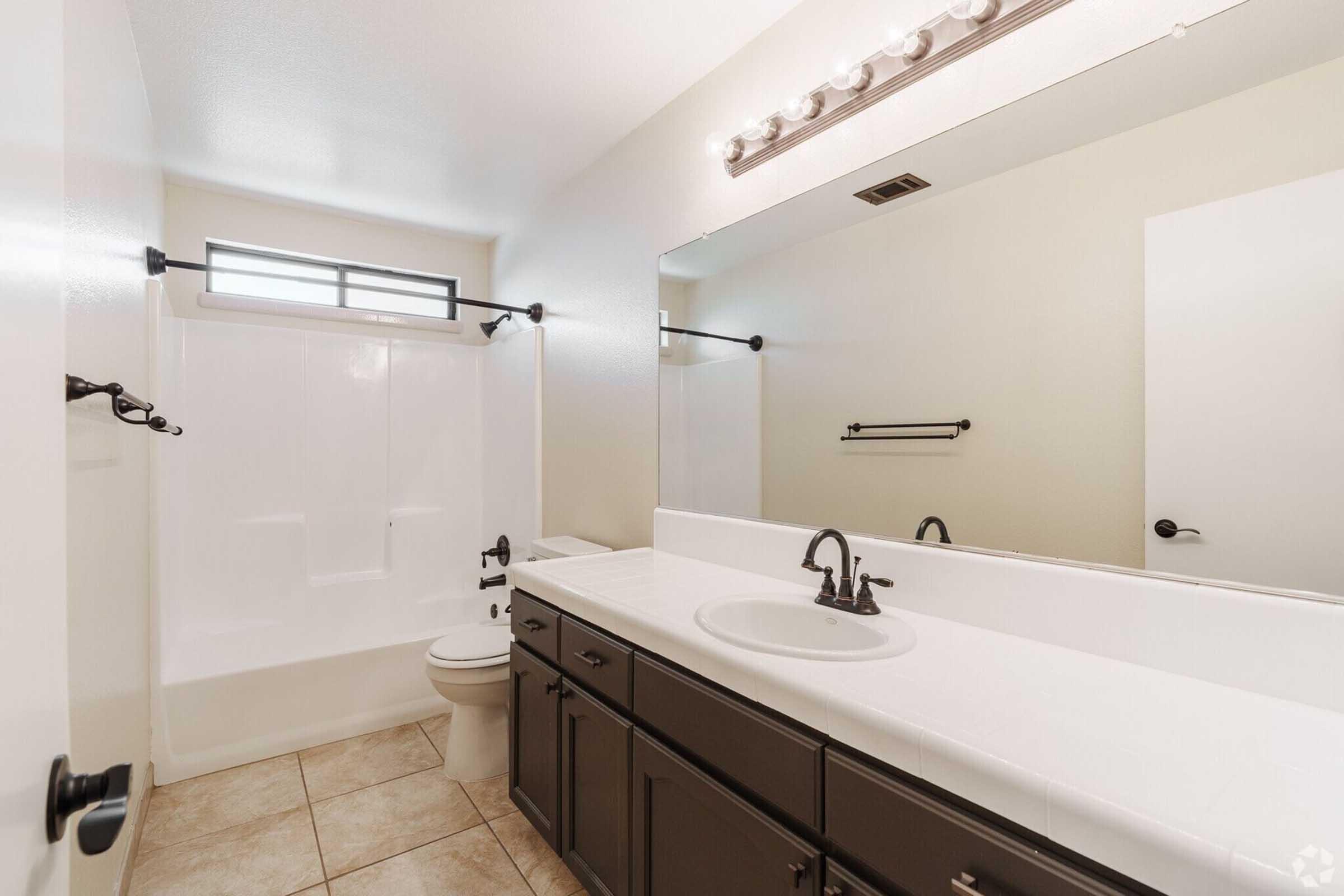 A bright and modern bathroom featuring a white bathtub with a shower, a sleek mirror above a double sink vanity with dark cabinetry, and light-colored tiled flooring. The room has natural light coming through a window and minimalistic fixtures for a clean and airy feel.