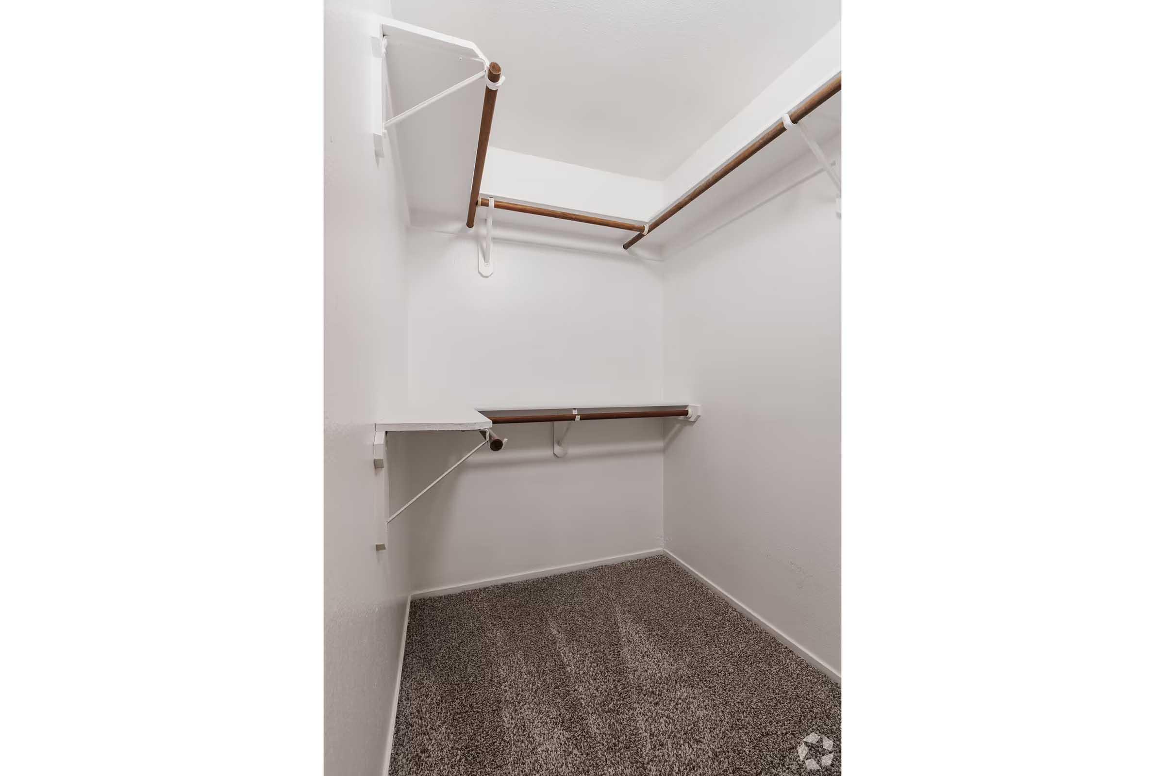 Empty closet with white walls, featuring two wooden shelves and two clothing rods mounted on the walls. The floor is covered in a soft, beige carpet, creating a clean and spacious appearance.