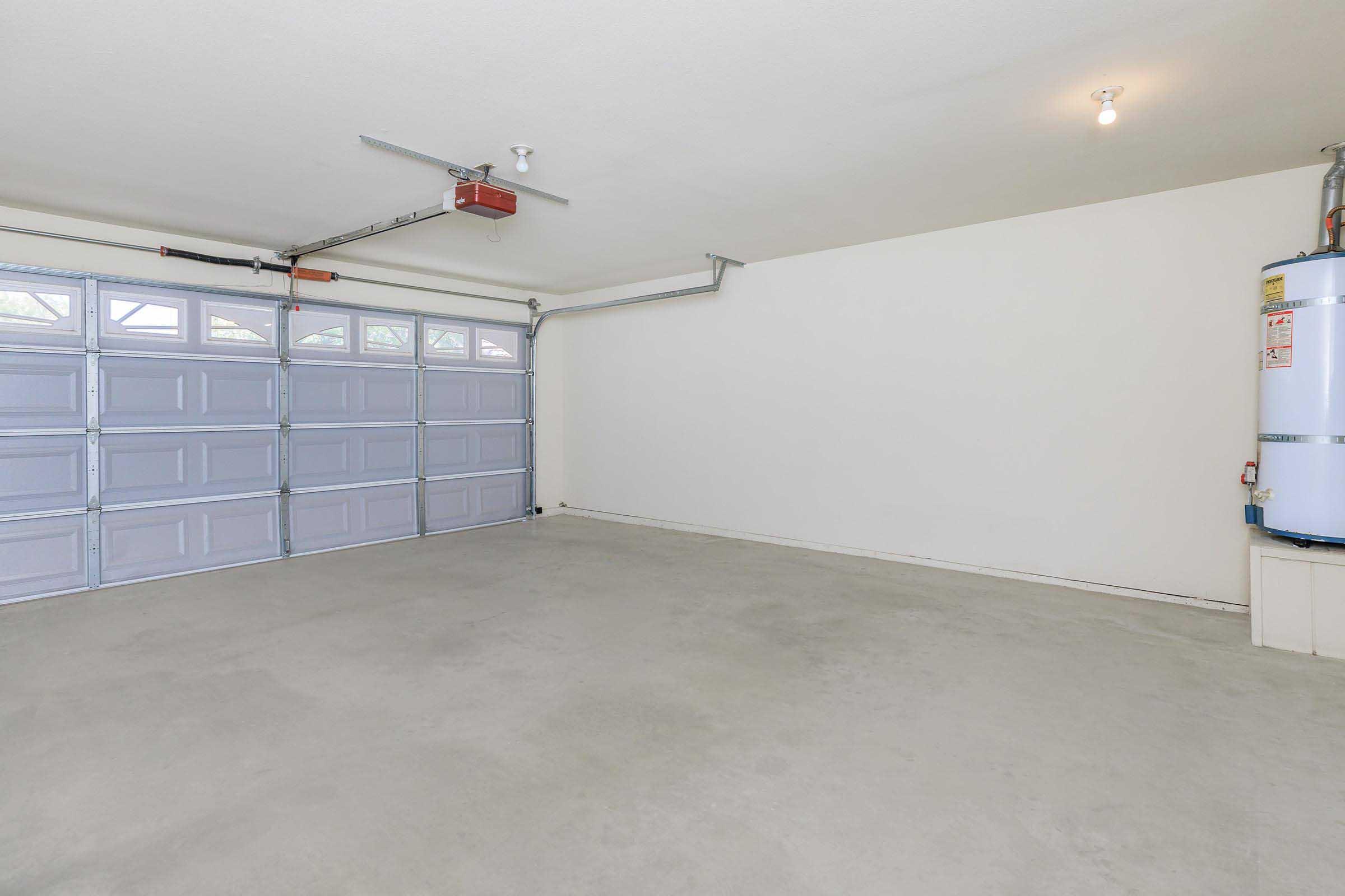 Empty garage with a concrete floor, featuring a garage door with four panels on the left side, a water heater mounted on the right wall, and a single overhead light fixture. Walls are painted a neutral color and the space is free of furniture or any other items.